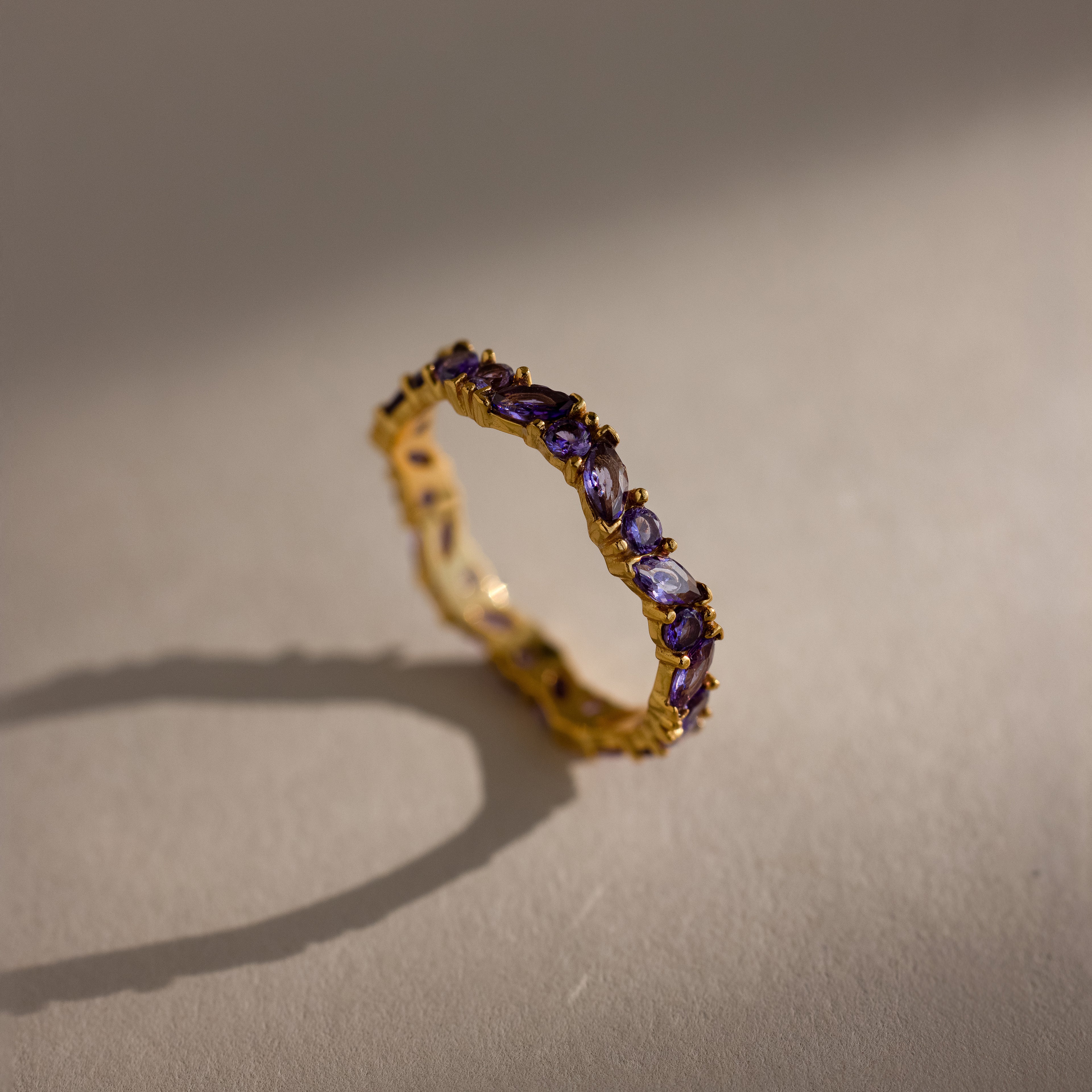 The Staggered Marquise Amethyst Ring casts a delicate shadow on a beige surface, its small purple gemstones sparkling softly.