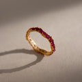 The Staggered Marquise Ruby Ring, featuring gold and red gemstones, rests on a beige surface and casts a soft shadow in the sunlight.