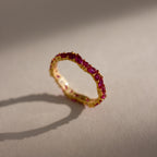 The Staggered Marquise Ruby Ring, featuring gold and red gemstones, rests on a beige surface and casts a soft shadow in the sunlight.