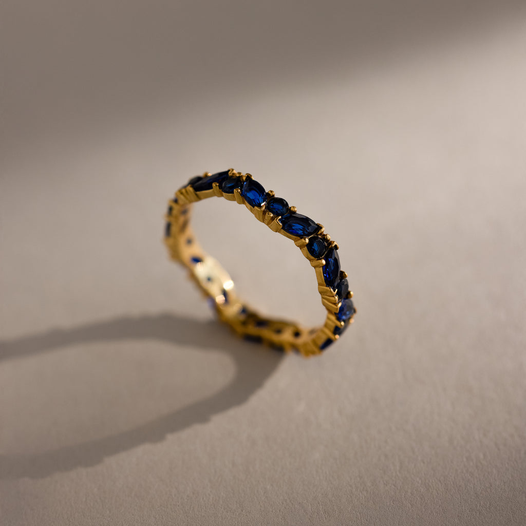 A Staggered Marquise Sapphire Ring—a gold eternity band set with multiple blue sapphires—casts a shadow on a beige surface.