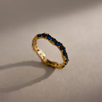 A Staggered Marquise Sapphire Ring—a gold eternity band set with multiple blue sapphires—casts a shadow on a beige surface.
