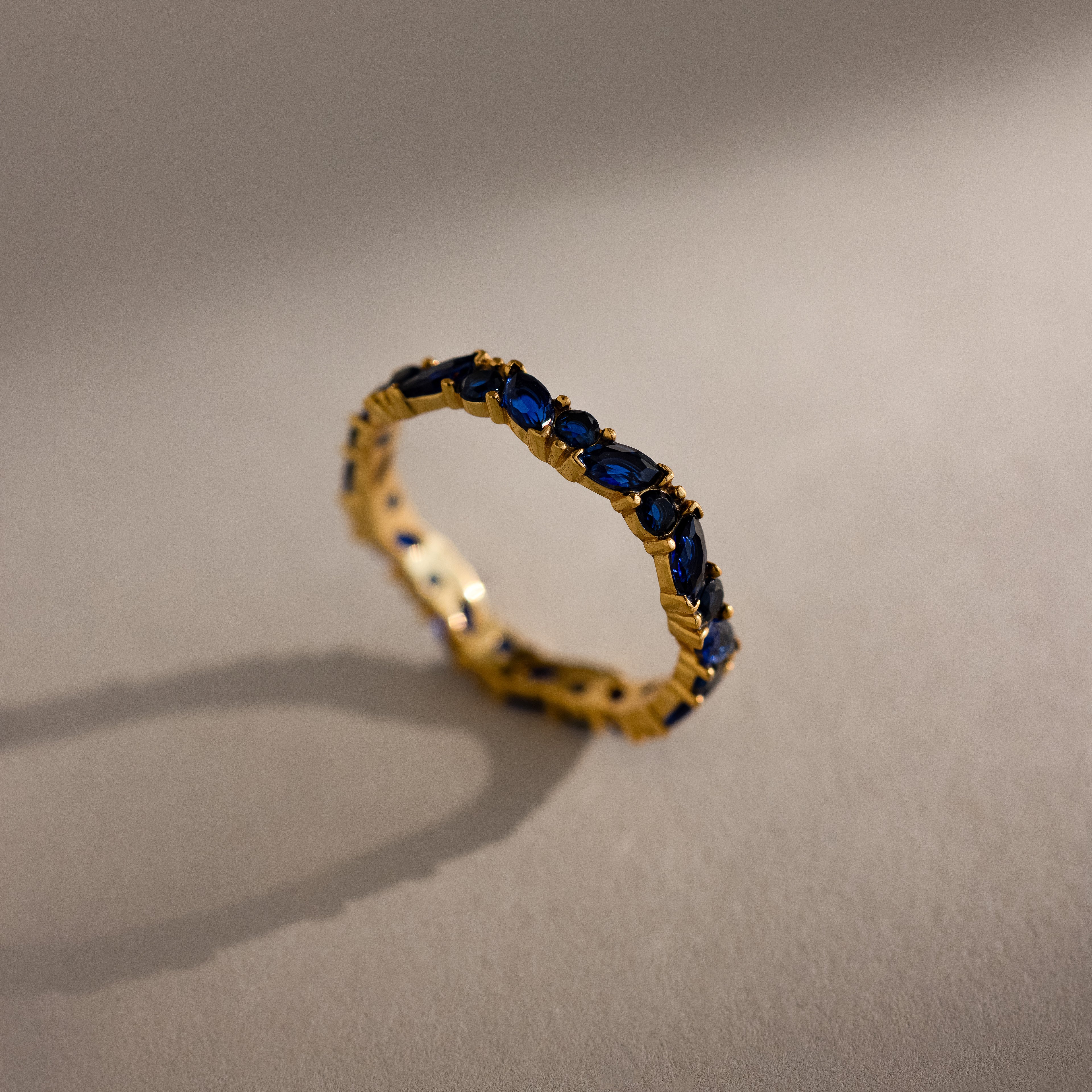 A Staggered Marquise Sapphire Ring—a gold eternity band set with multiple blue sapphires—casts a shadow on a beige surface.