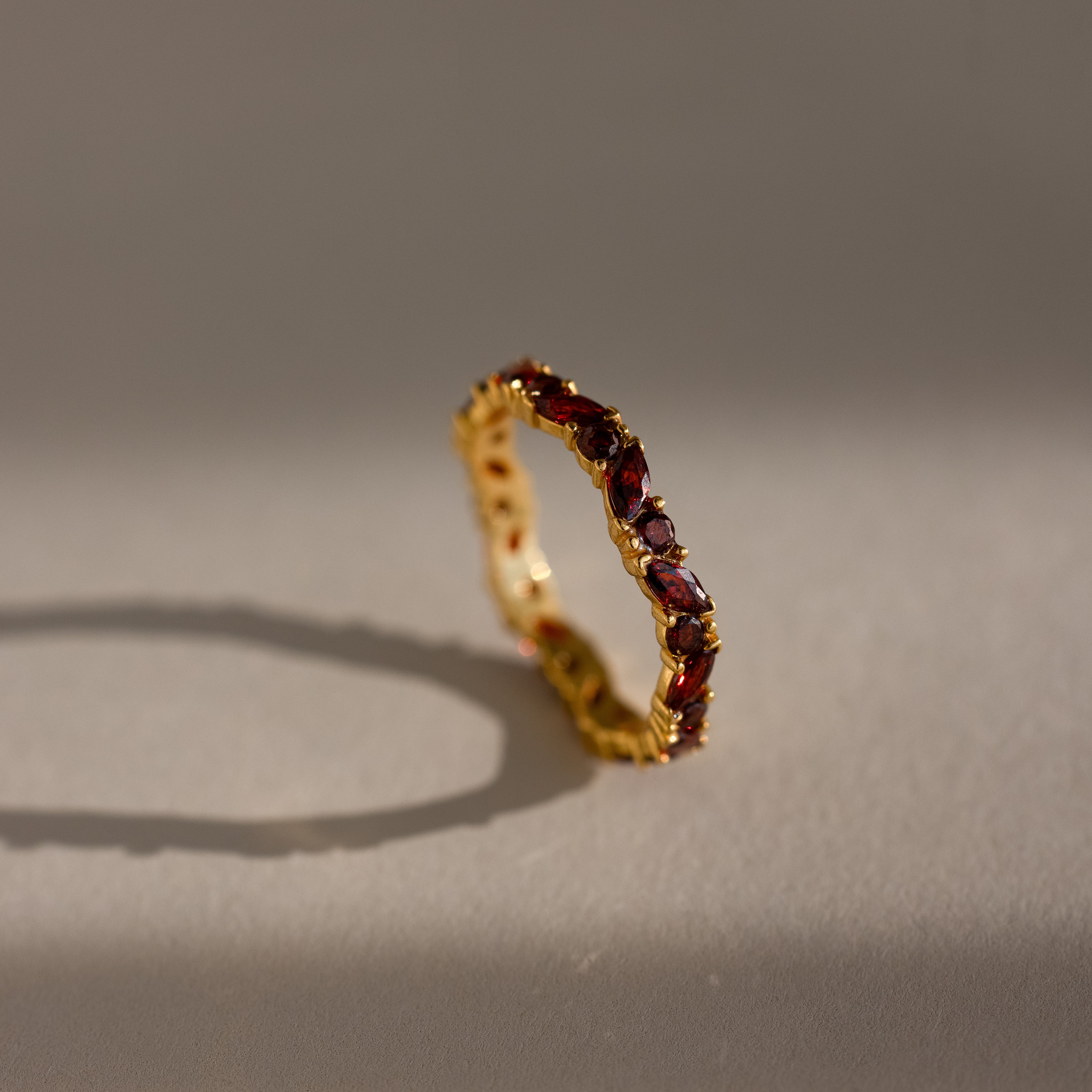 The Staggered Marquise Garnet Ring shines with red gemstones, casting a shadow on a softly lit neutral surface.