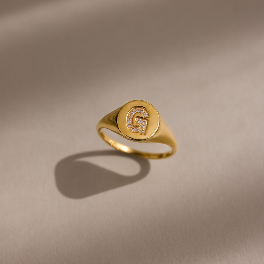 The Pave Initial Signet Ring in 18K Gold features a crystal-studded initial G that sparkles with small diamonds on a neutral background, making it the perfect statement ring.