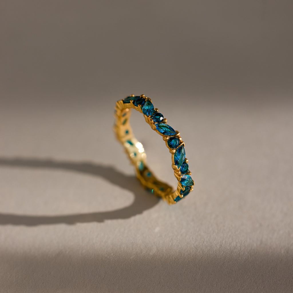 The Staggered Marquise Blue Zircon Ring features gleaming marquise-cut blue zircons set in gold, standing upright on a neutral surface and casting soft shadows—an elegant eternity band choice.