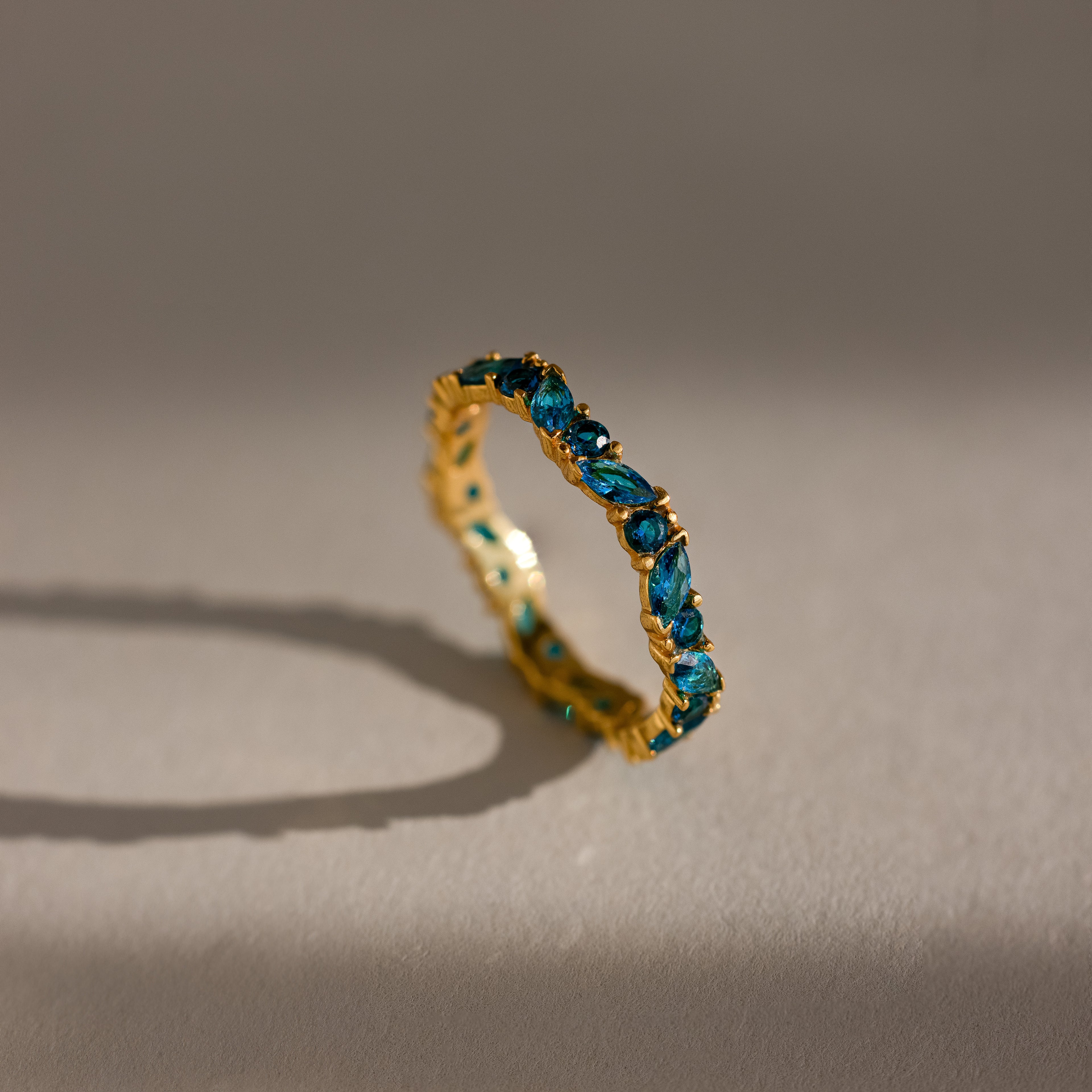The Staggered Marquise Blue Zircon Ring features gleaming marquise-cut blue zircons set in gold, standing upright on a neutral surface and casting soft shadows—an elegant eternity band choice.