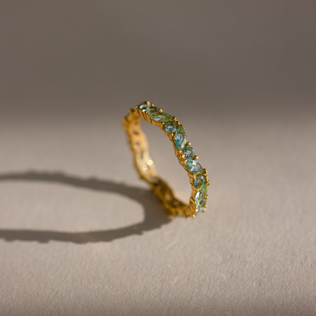 The Staggered Marquise Aquamarine Ring features gold and blue marquise gemstones in an eternity band, casting a shadow on a beige surface.