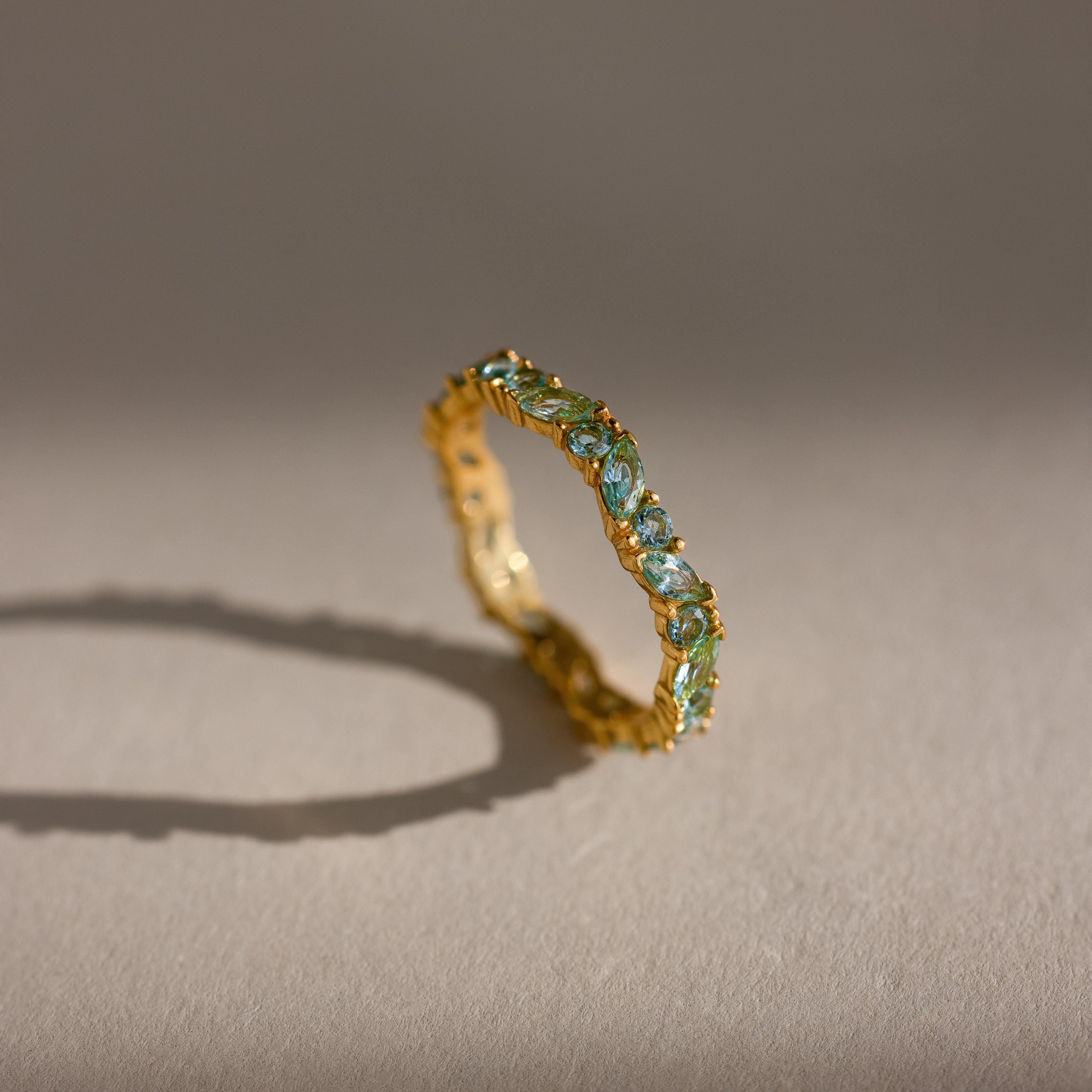 The Staggered Marquise Aquamarine Ring features gold and blue marquise gemstones in an eternity band, casting a shadow on a beige surface.