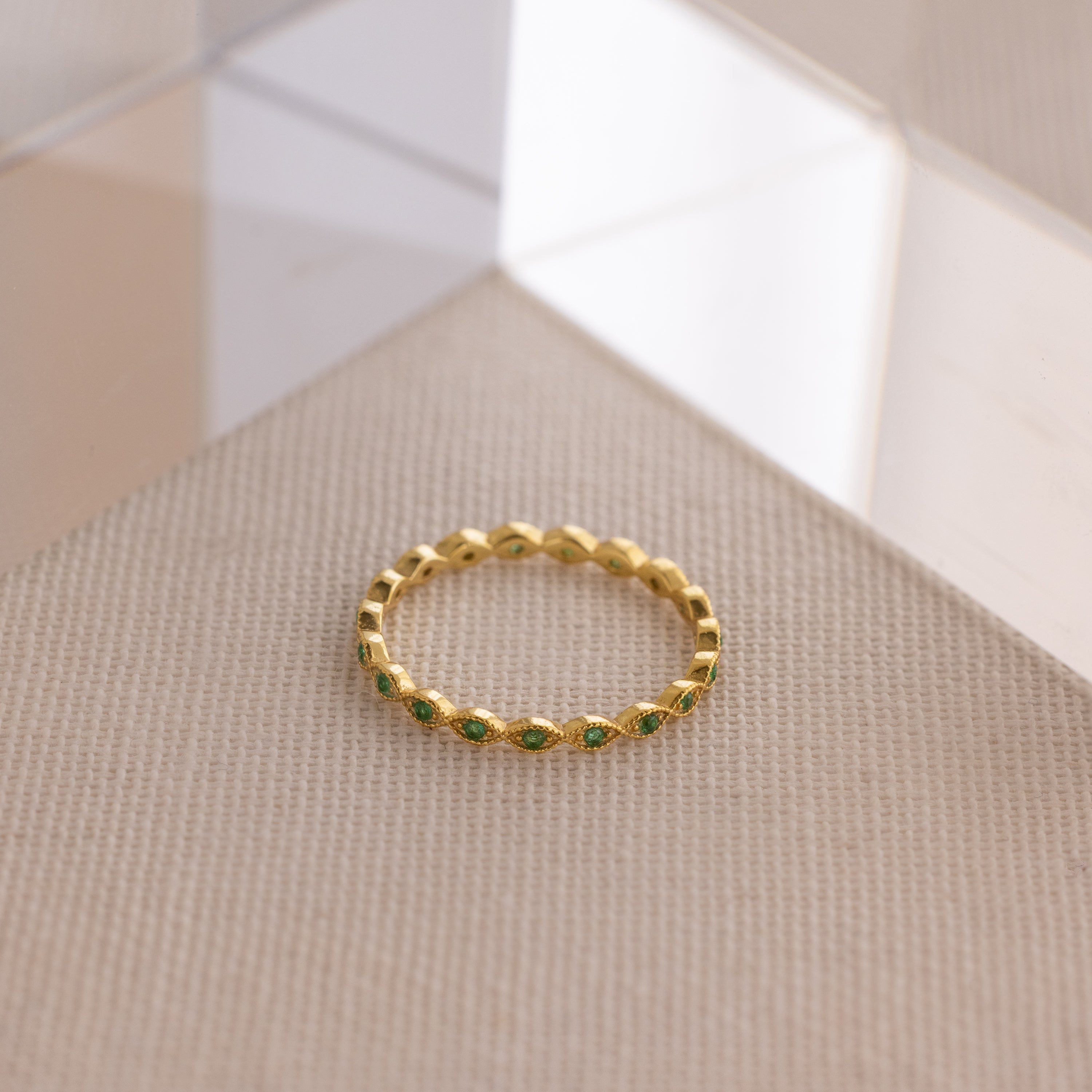 The Dainty Marquise Emerald Ring, featuring green gemstones with an Art Deco vibe, rests on a textured beige surface near a clear acrylic block.