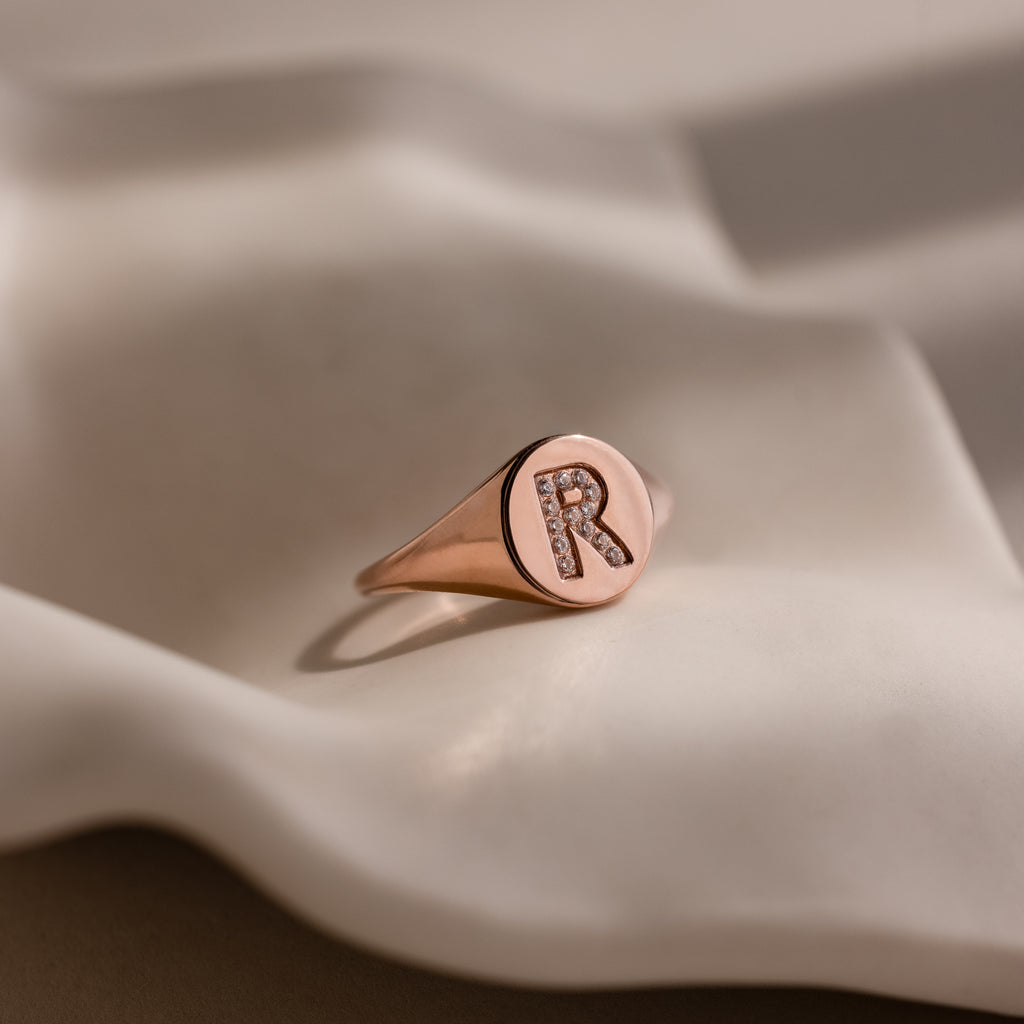 The Pave Initial Signet Ring in Rose Gold, adorned with a diamond-studded letter R, is showcased on a wavy, light-colored surface.