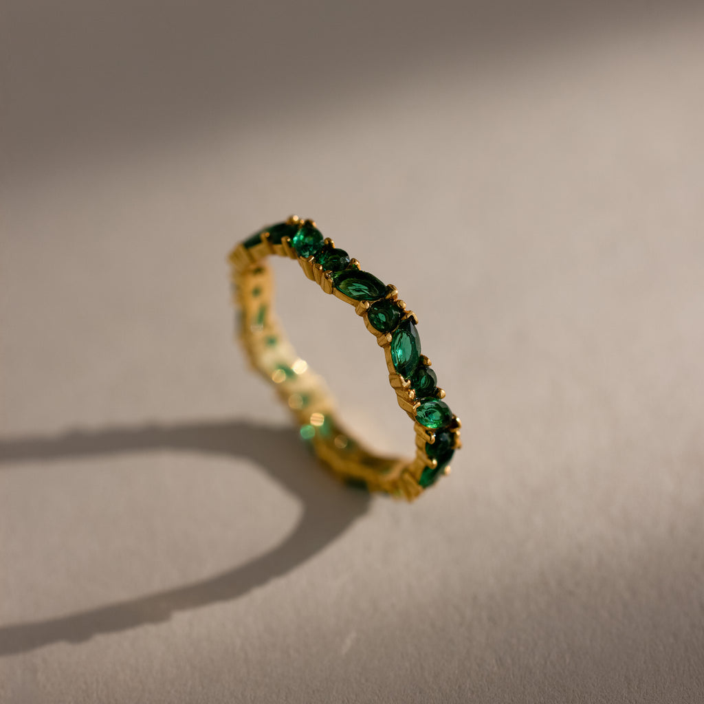 The Staggered Marquise Emerald Ring, a gold eternity band set with green emeralds, casts a shadow on a neutral surface.