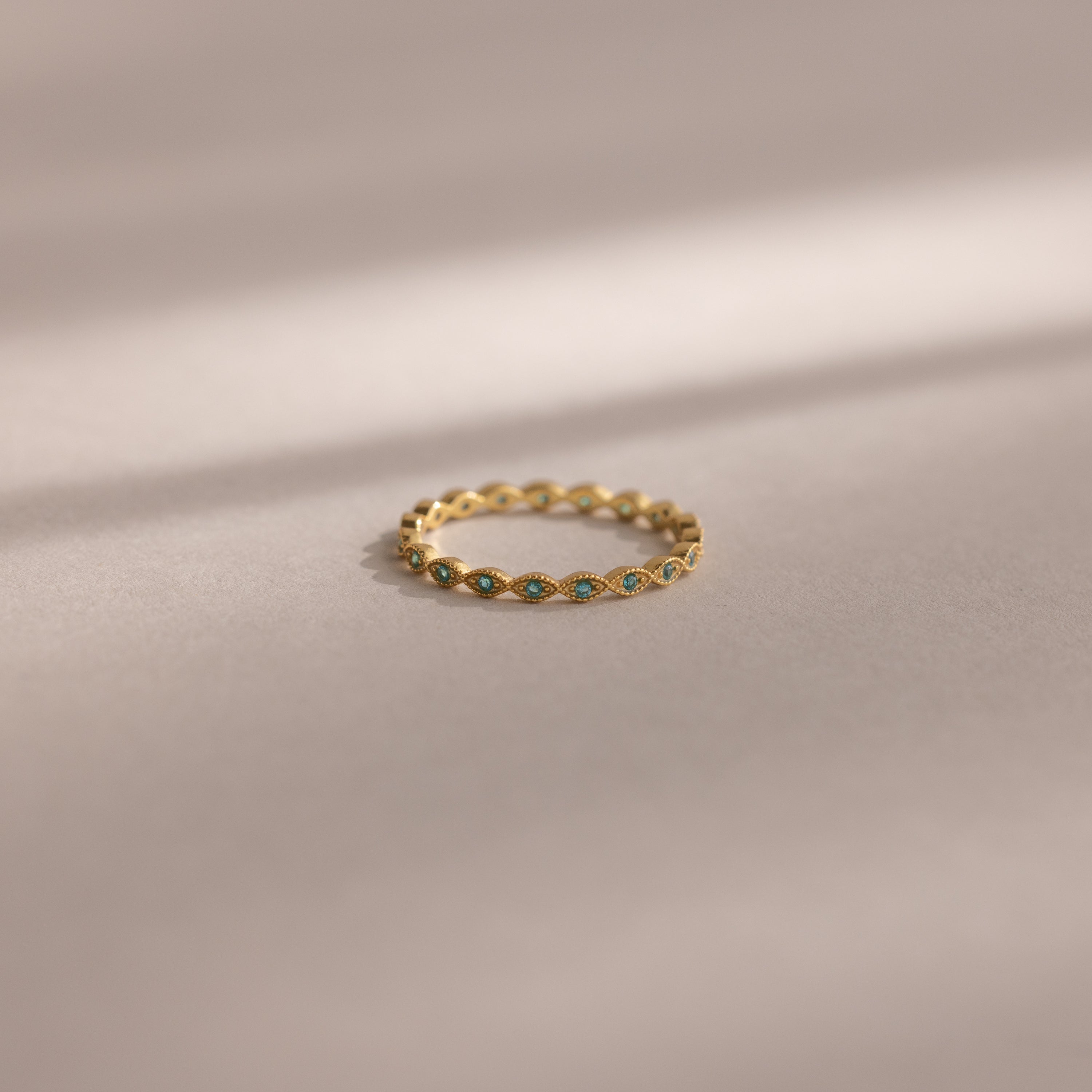 The Dainty Marquise Blue Zircon Ring, a delicate Art Deco-inspired gold piece with a shimmering blue zircon, rests on a soft beige surface with gentle shadow lines.