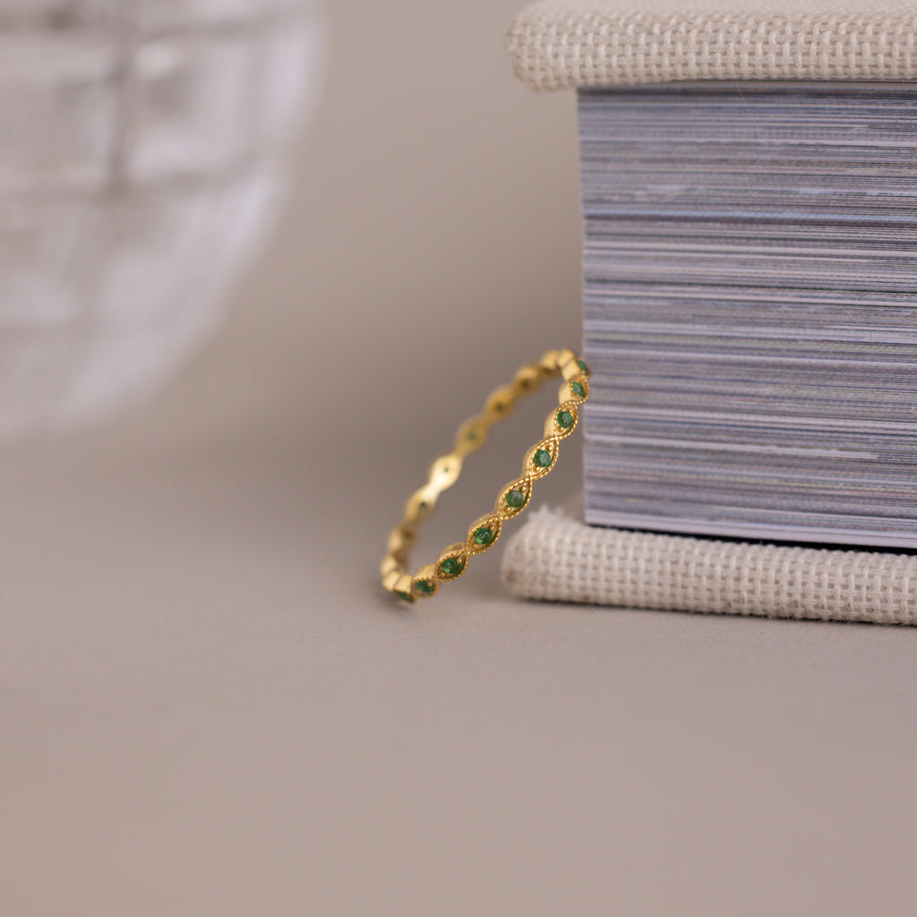 The Dainty Marquise Emerald Ring stands upright against a textured box on a neutral surface, showcasing its delicate design and hint of Art Deco charm.