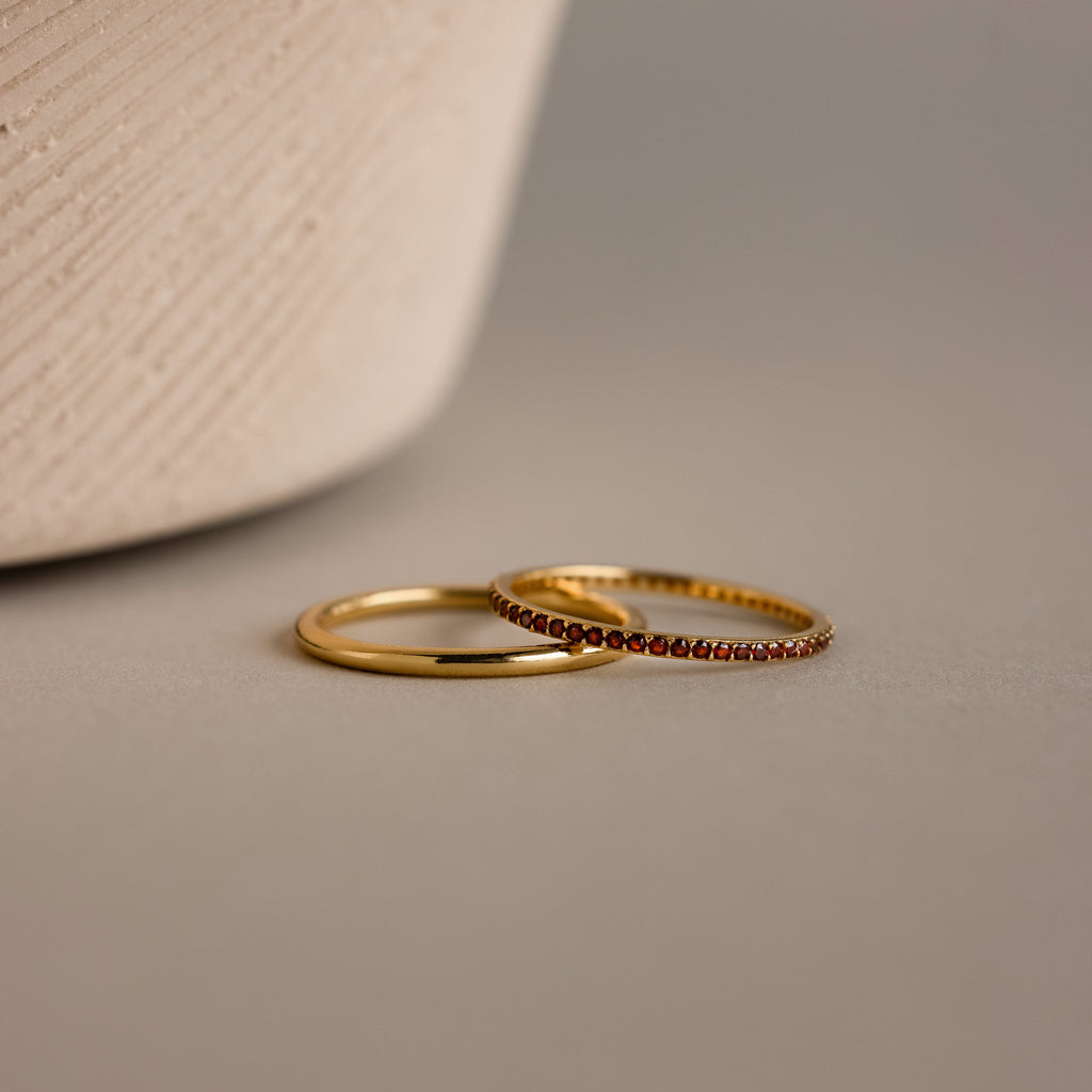 The Birthstone Tennis Ring Set in Garnet (January) features a classic gold band and a gemstone eternity ring with small red stones, both elegantly displayed on a neutral surface.
