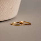 The Birthstone Tennis Ring Set in Garnet (January) features a classic gold band and a gemstone eternity ring with small red stones, both elegantly displayed on a neutral surface.