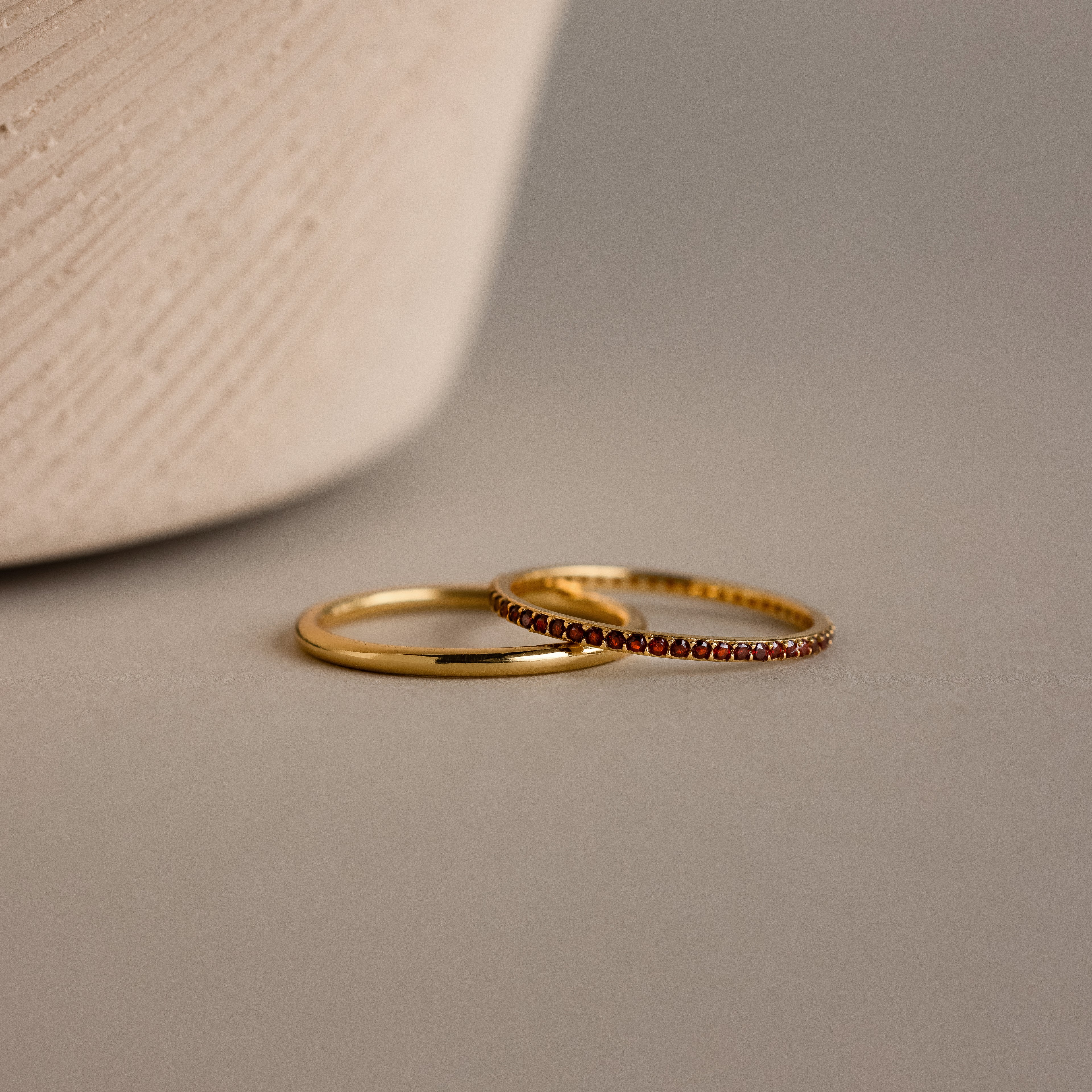 The Birthstone Tennis Ring Set in Garnet (January) features a classic gold band and a gemstone eternity ring with small red stones, both elegantly displayed on a neutral surface.