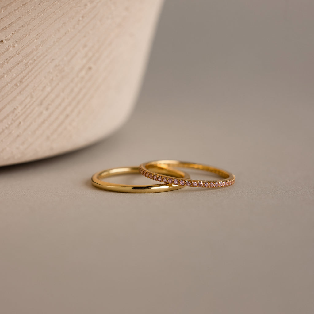 The Pink Tourmaline Tennis Ring Set, featuring a classic gold band and a gemstone eternity ring with sparkling tourmalines, is displayed on a neutral surface next to a textured vase.