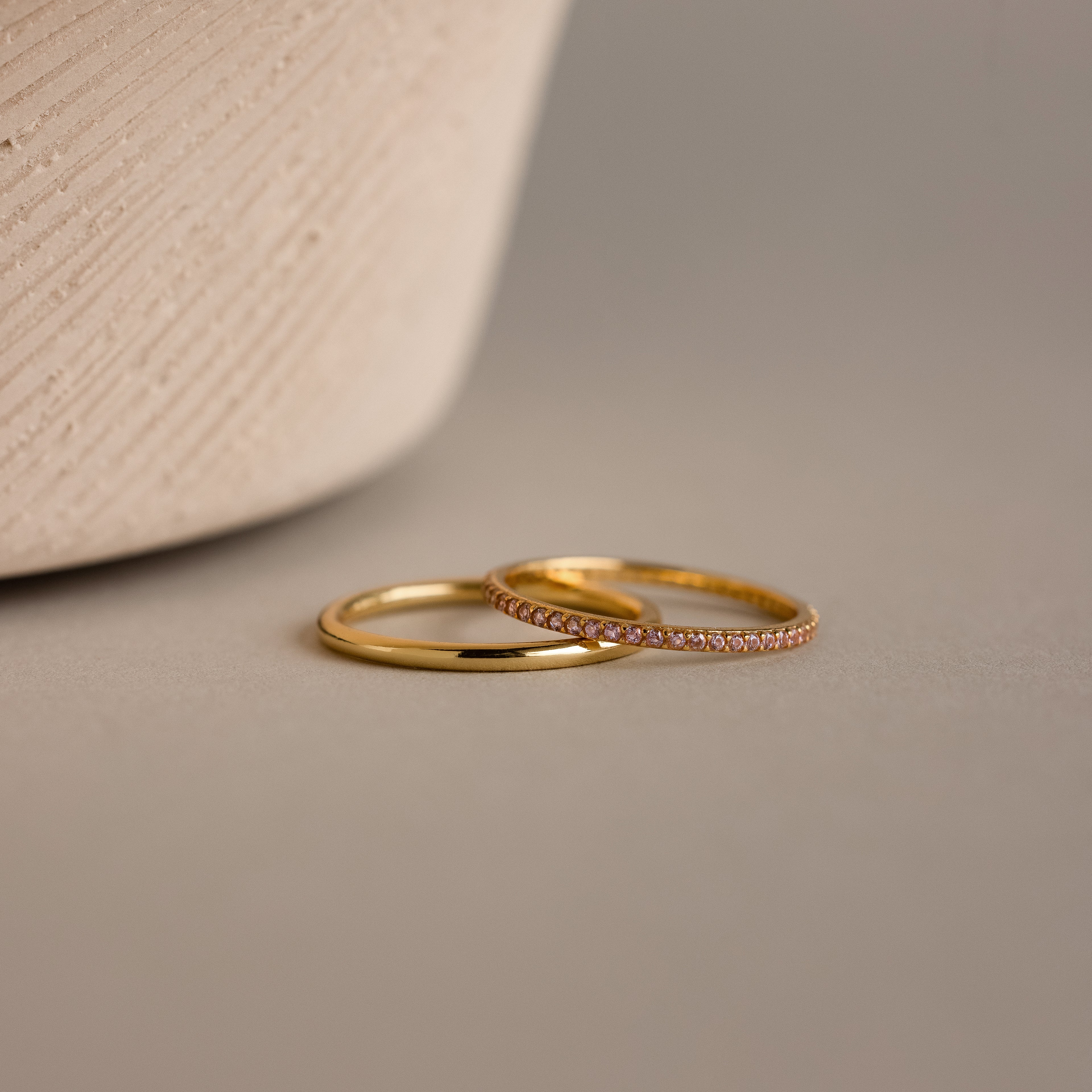 The Pink Tourmaline Tennis Ring Set, featuring a classic gold band and a gemstone eternity ring with sparkling tourmalines, is displayed on a neutral surface next to a textured vase.