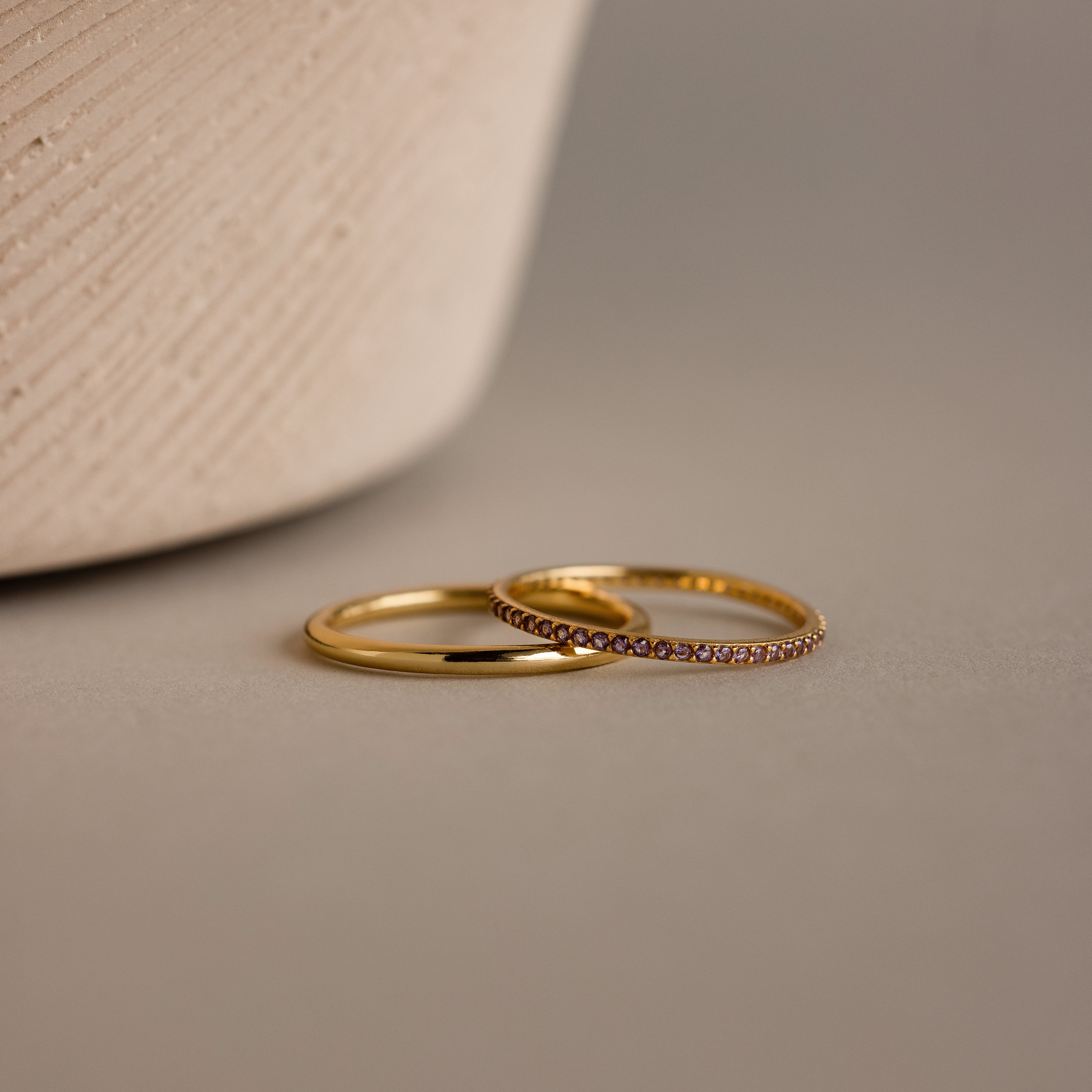 Close-up of a delicate gold band ring set with a continuous row of sparkling alexandrite gemstones, showcasing their soft violet-lavender hue. The ring rests on a sculptural, ribbed ceramic surface in a warm neutral tone, emphasizing the contrast between the luminous gold finish and the subtle shimmer of the birthstones.
