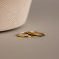 The Sapphire Tennis Ring Set, featuring one plain gold ring and one with subtle blue gemstones, rests elegantly on a neutral surface.