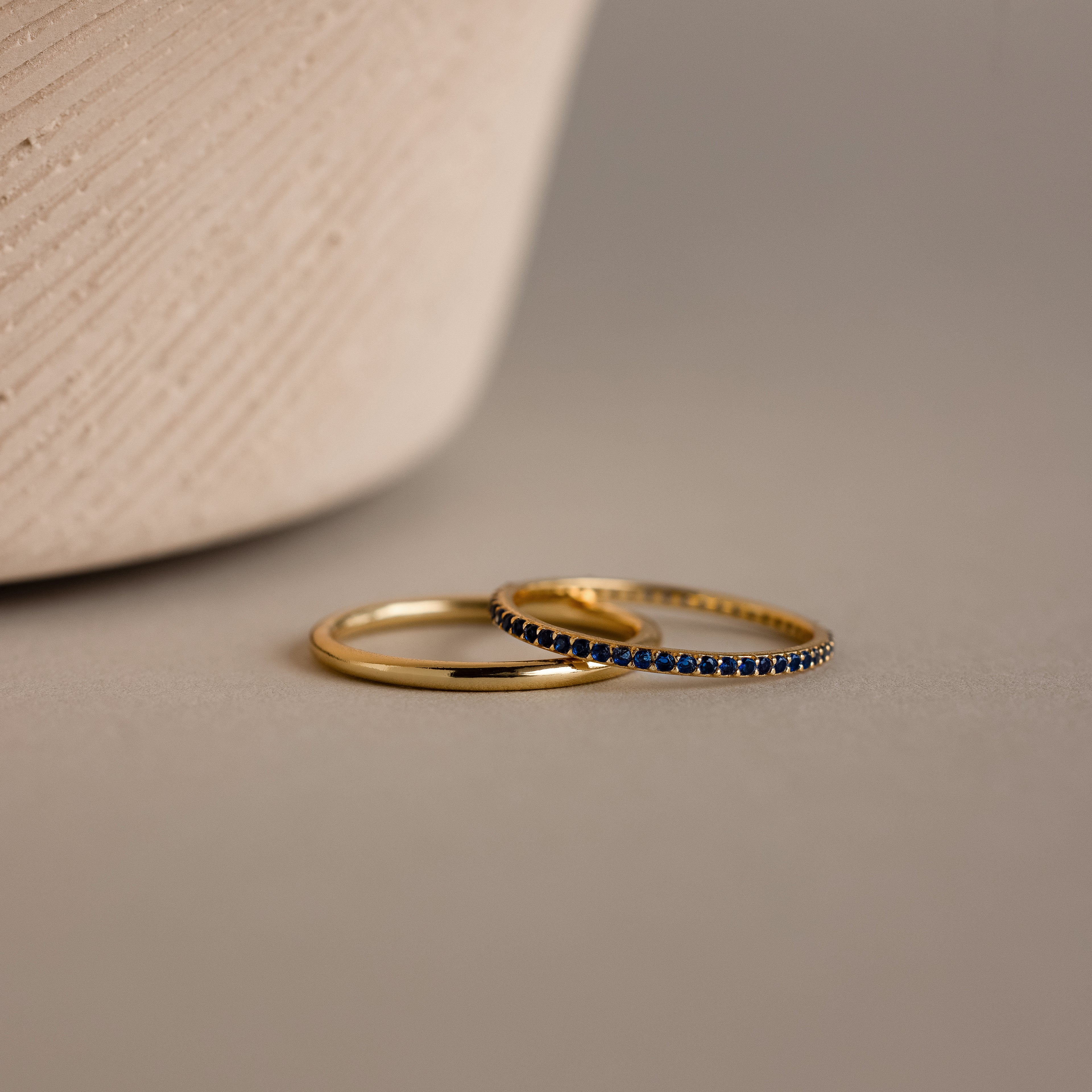 The Sapphire Tennis Ring Set, featuring one plain gold ring and one with subtle blue gemstones, rests elegantly on a neutral surface.