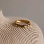 A close-up shot of a thin gold band inlaid with amethyst-hued stones, resting elegantly on a textured, cream-toned ceramic background.