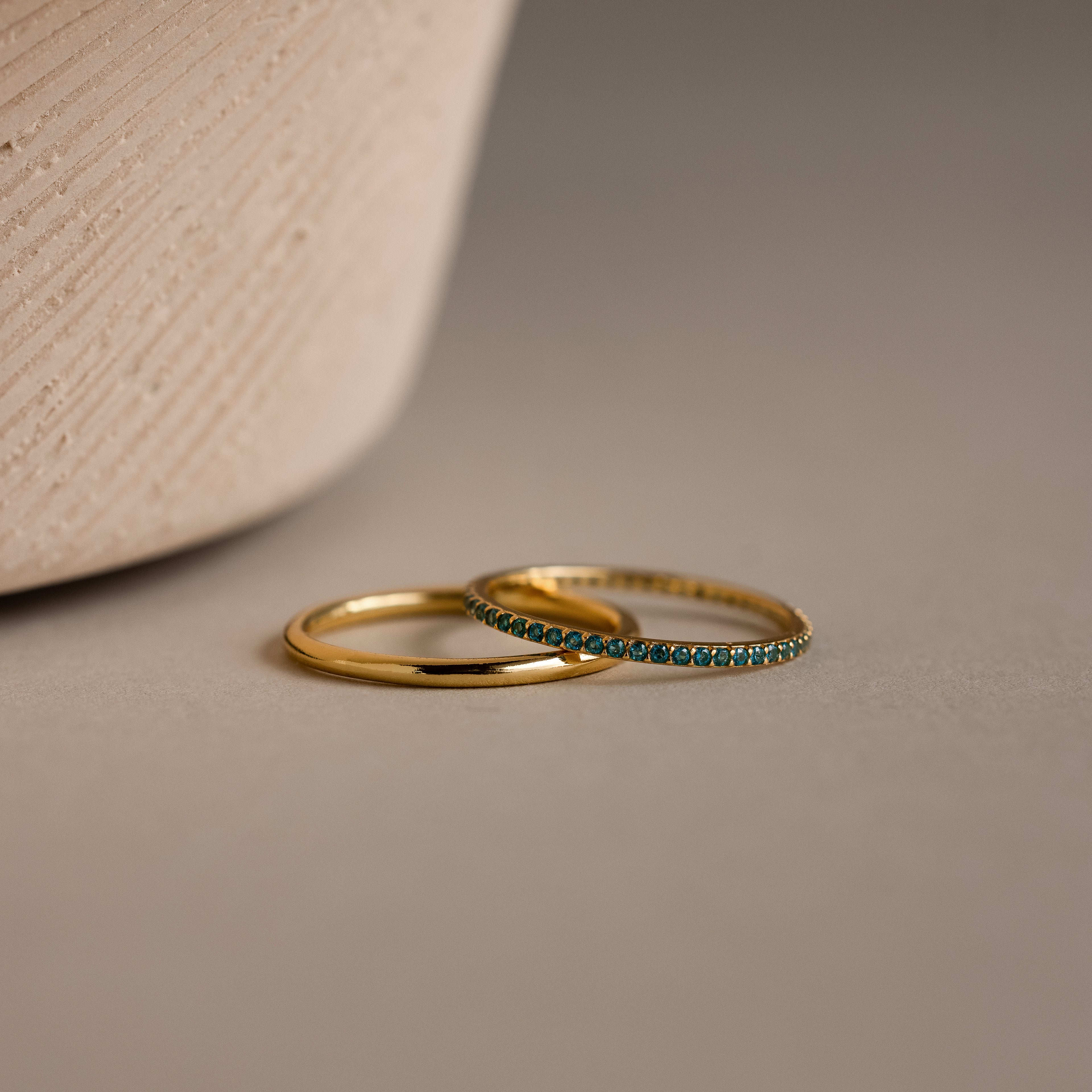The Blue Zircon Tennis Ring Set, featuring one plain gold ring and another adorned with blue zircon stones in an eternity style, rests on a neutral surface beside a textured pot.