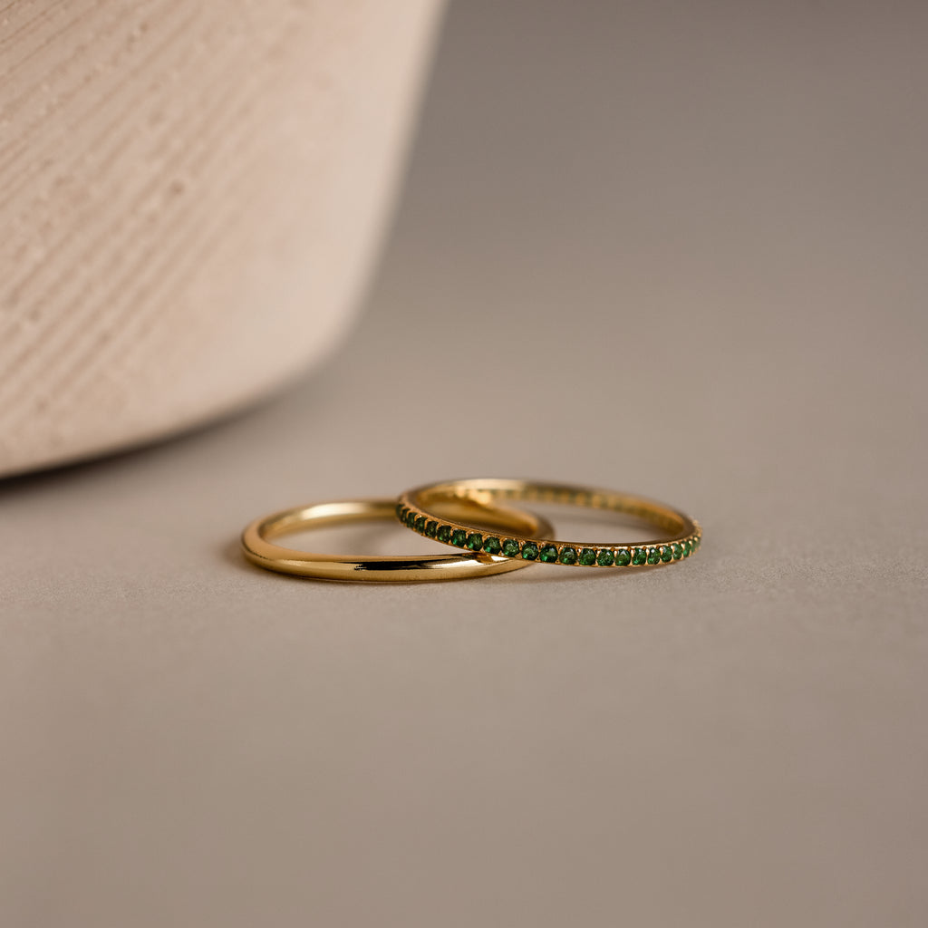 The Birthstone Tennis Ring Set in Emerald/May features two delicate gold rings—a sleek band and a gemstone eternity ring with small green stones—resting elegantly on a neutral surface.