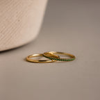 The Birthstone Tennis Ring Set in Emerald/May features two delicate gold rings—a sleek band and a gemstone eternity ring with small green stones—resting elegantly on a neutral surface.