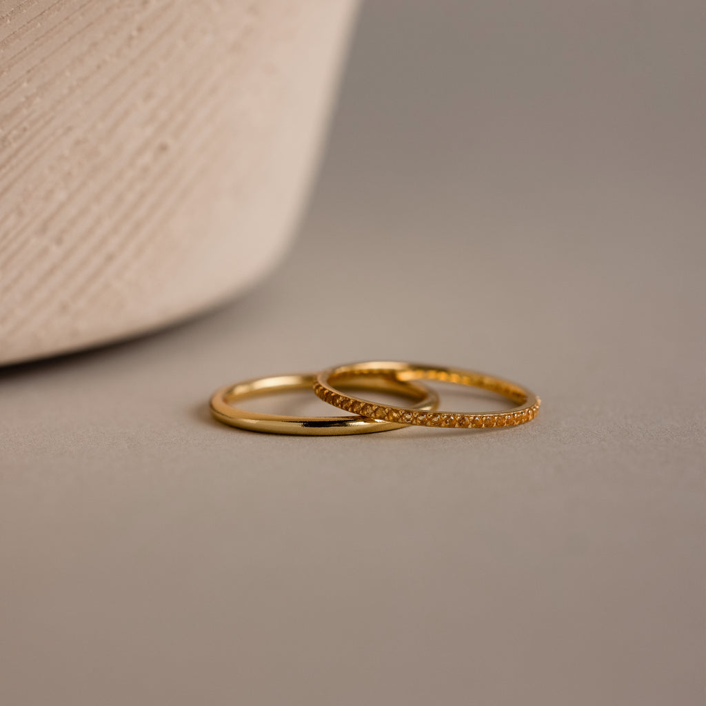 The Topaz Tennis Ring Set features two delicate gold rings—one simple band and one adorned with sparkling stones—resting on a smooth beige surface, ideal for stacking with your favorite birthstone ring.
