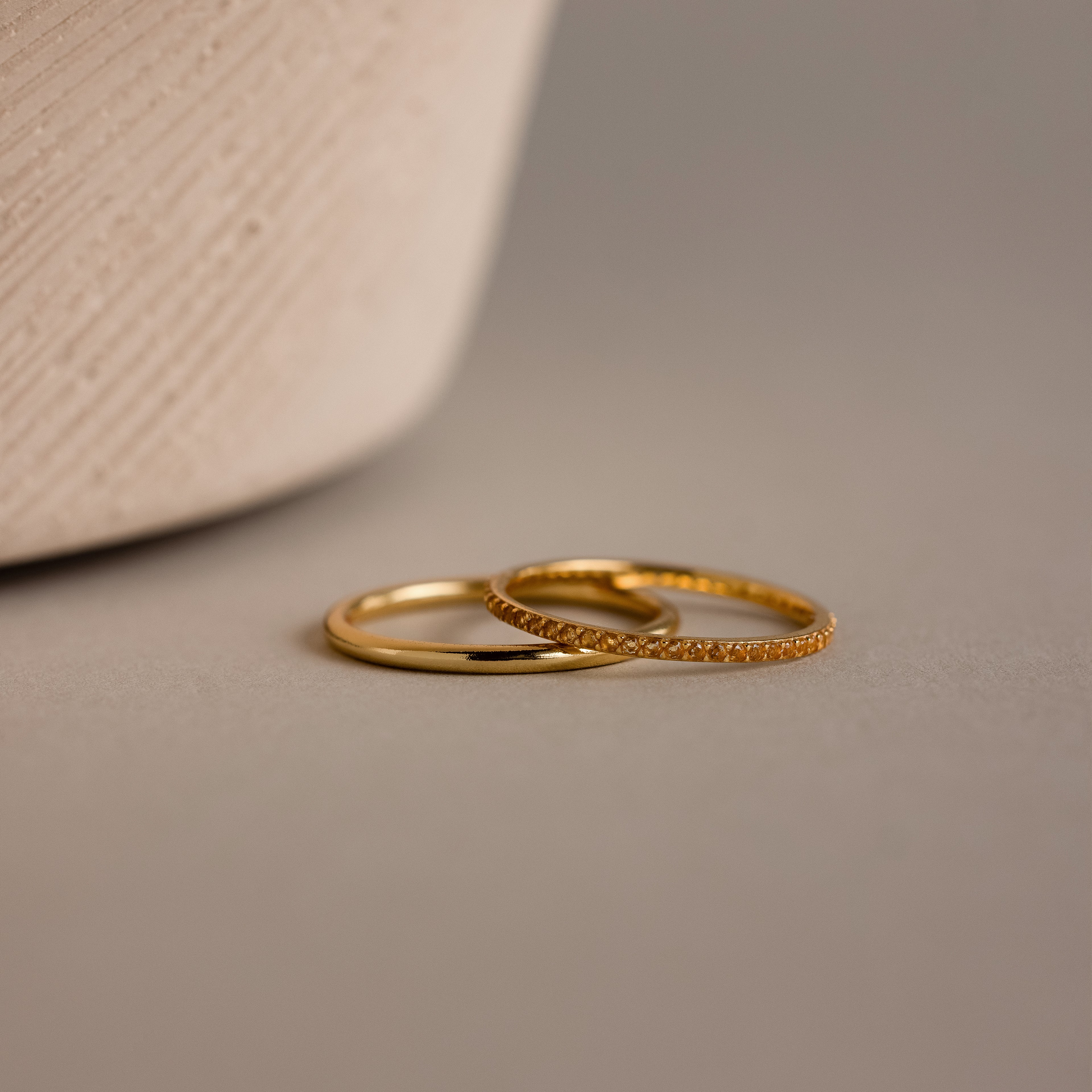 The Topaz Tennis Ring Set features two delicate gold rings—one simple band and one adorned with sparkling stones—resting on a smooth beige surface, ideal for stacking with your favorite birthstone ring.