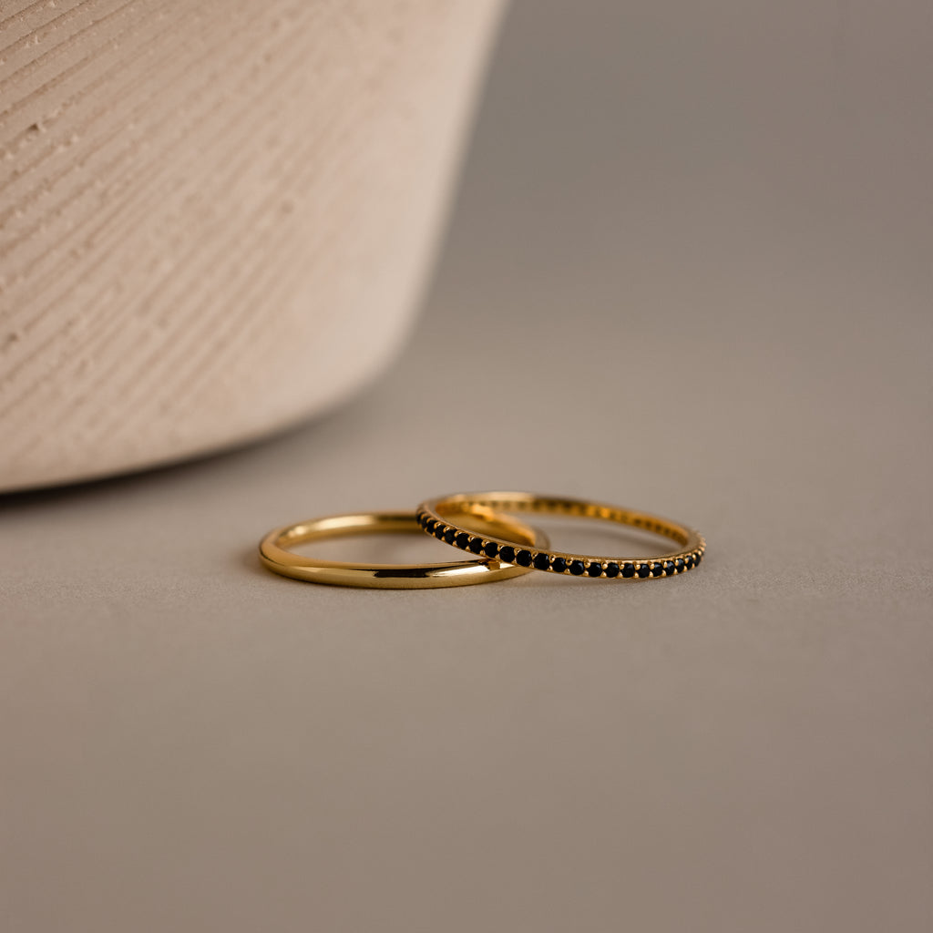 The Onyx Tennis Ring Set—featuring a classic gold band and a black gemstone eternity ring—rests on a smooth beige surface near a textured vase.