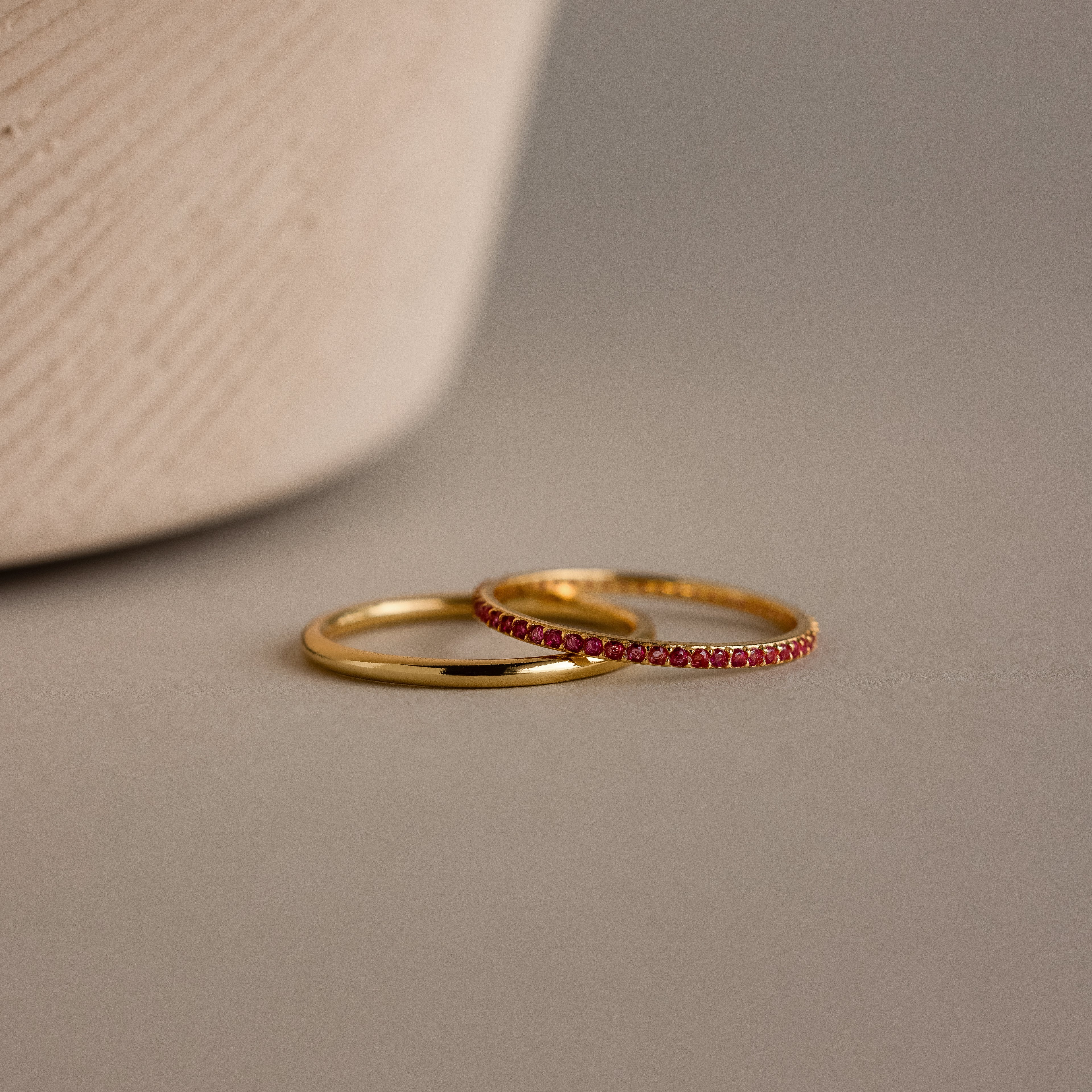 Two delicate gold rings, one plain and the other the Ruby Tennis Ring Set adorned with small red stones, rest on a neutral surface.