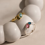 The Toi et Moi Heart Birthstone Ring in Rose Gold is displayed on a smooth, white, rounded surface.