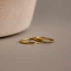The Peridot Tennis Ring Set, featuring one simple gold band and one gold eternity ring with small peridot stones, is displayed on a neutral surface beside a textured object.