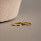 The Amethyst Tennis Ring Set, featuring a plain gold ring and an amethyst-studded band with small purple stones, rests on a neutral surface beside a textured object.
