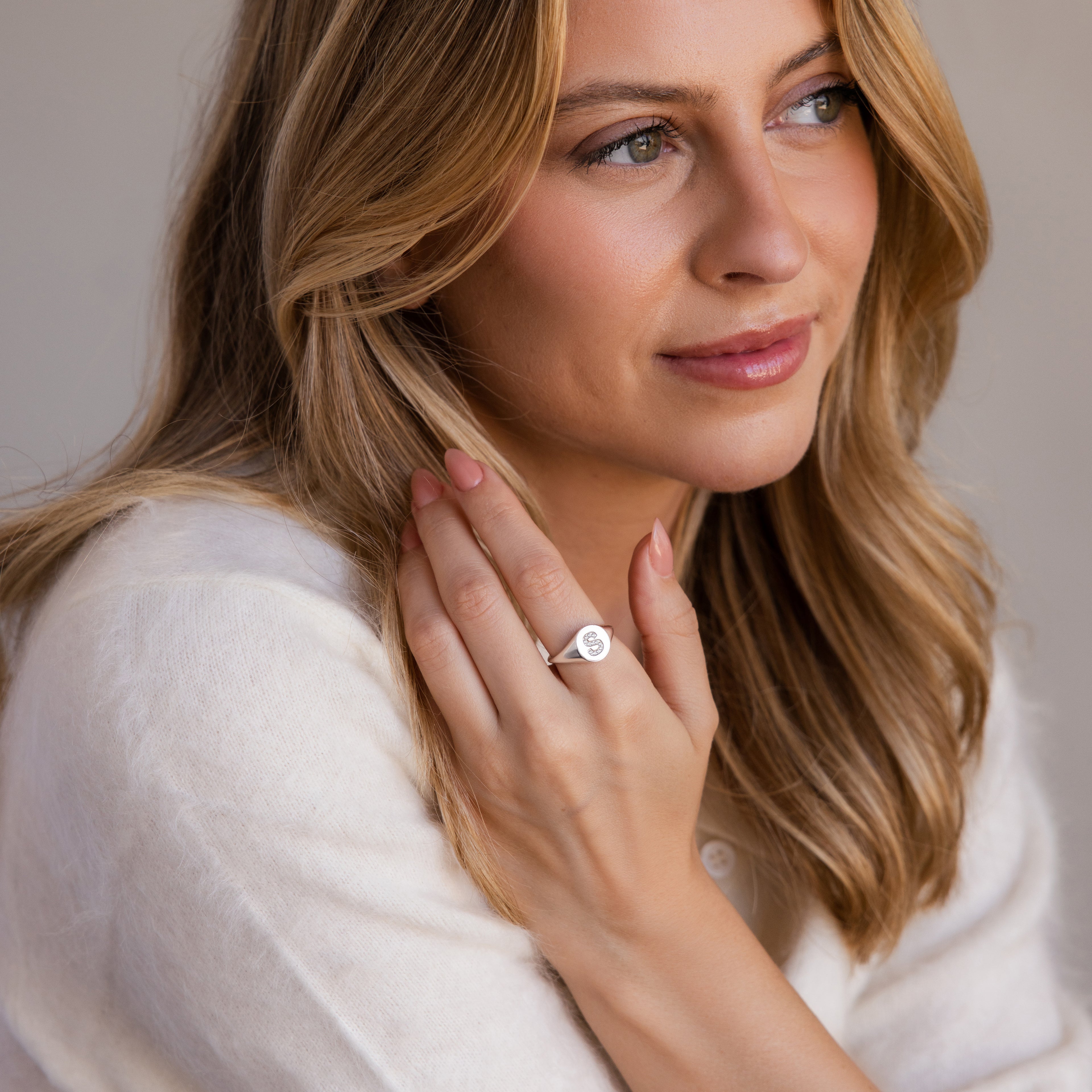Woman with wavy blonde hair wearing a white top and the Pave Initial Signet Ring, looking thoughtfully to the side.