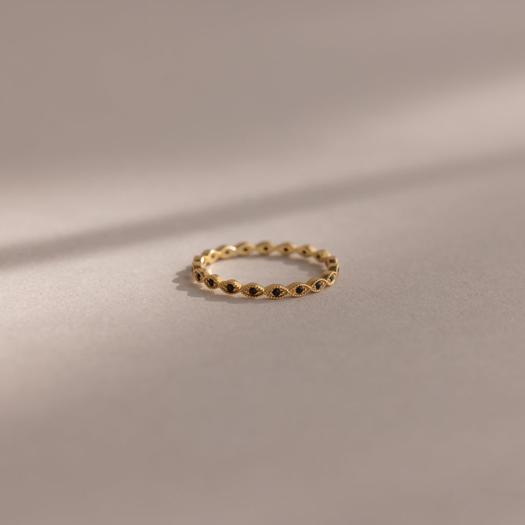The Dainty Marquise Onyx Ring, a delicate gold Art Deco design with small, evenly spaced round accents, rests on a soft beige surface with gentle shadows.