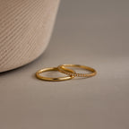 The Birthstone Tennis Ring Set in Diamond / April features two delicate gold rings—one a simple band, the other an eternity ring with sparkling stones—displayed on a neutral surface beside a textured vase.
