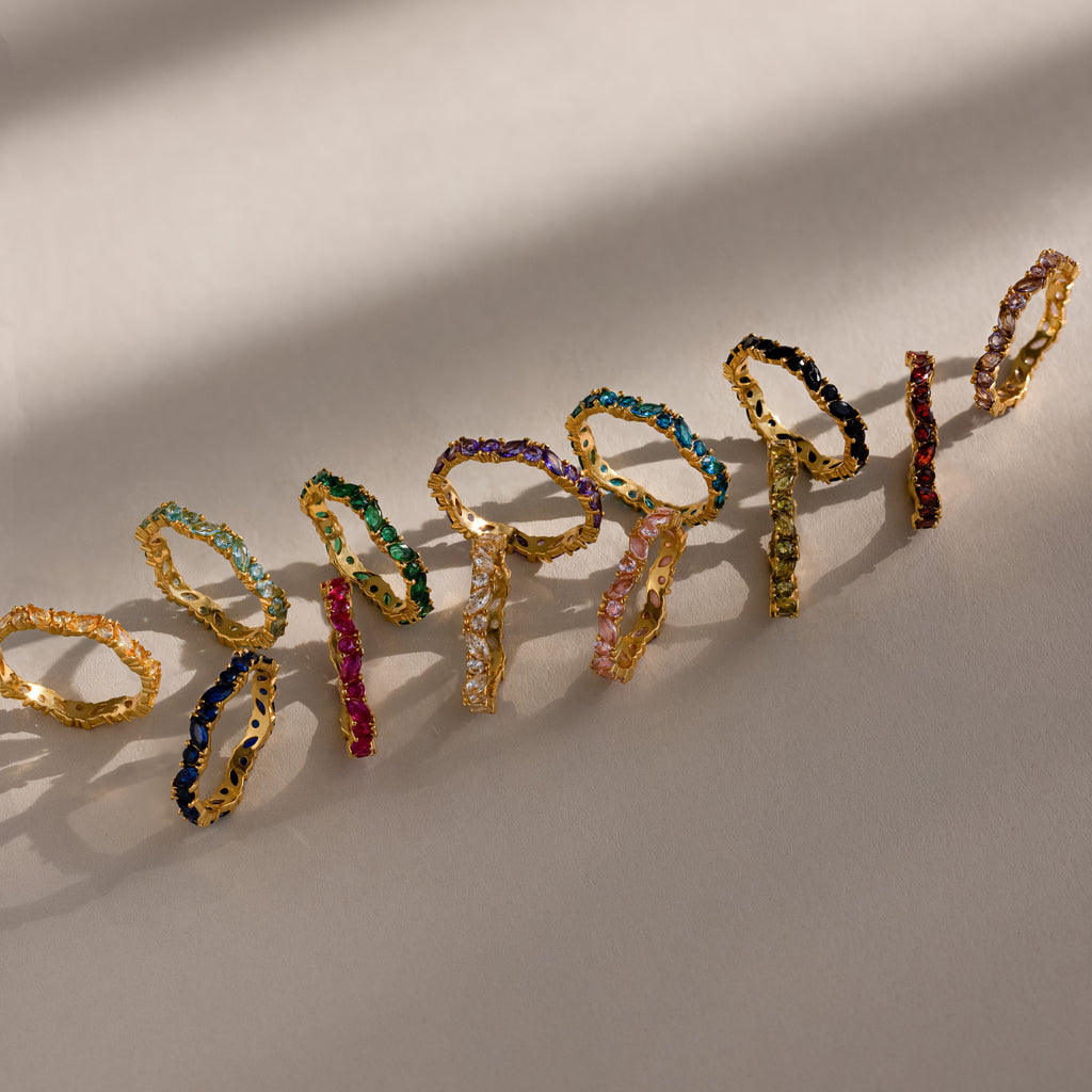 The Staggered Marquise Birthstone Ring features colorful gemstones set in gold, arranged in a row on a beige surface and casting soft shadows.