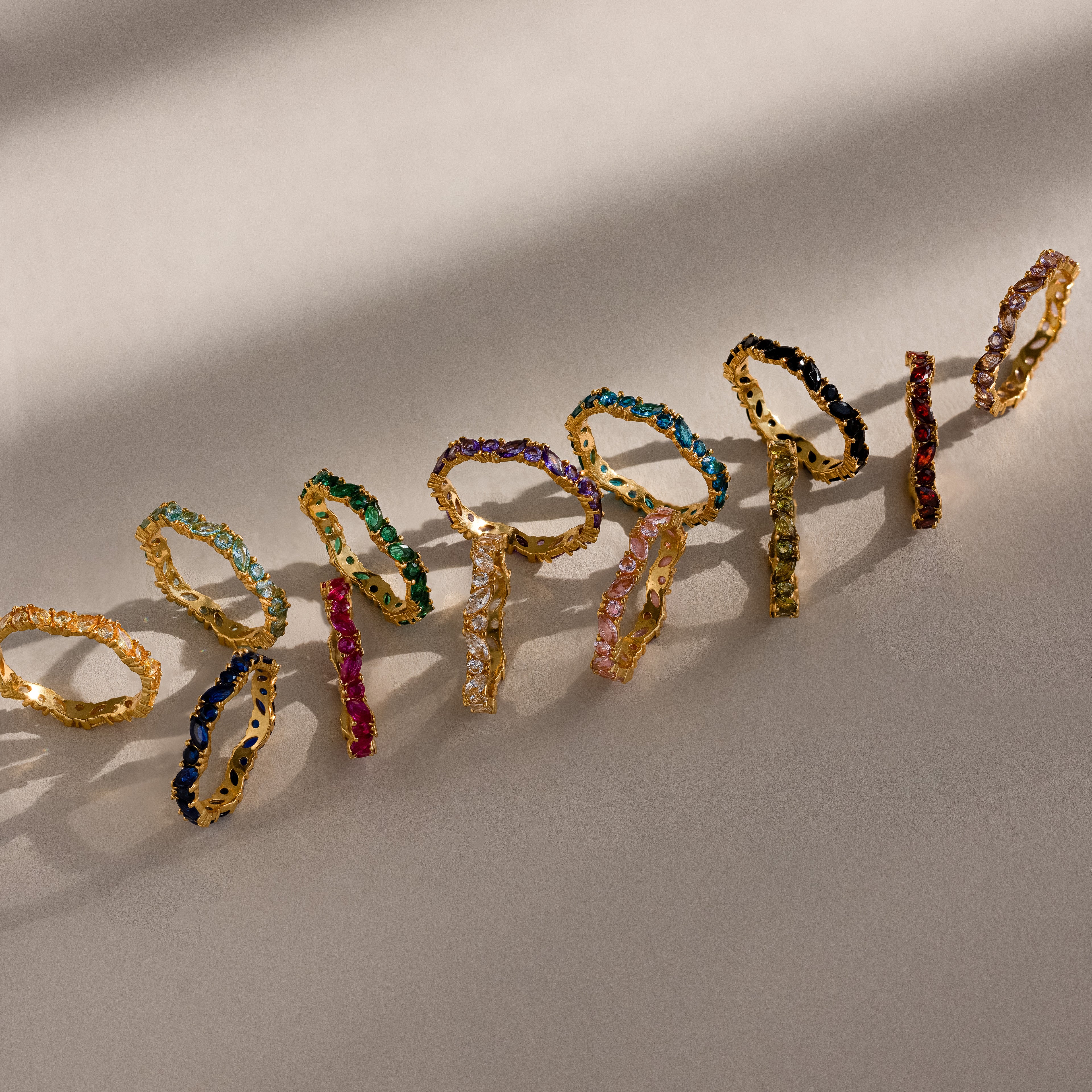The Staggered Marquise Birthstone Ring features colorful gemstones set in gold, arranged in a row on a beige surface and casting soft shadows.