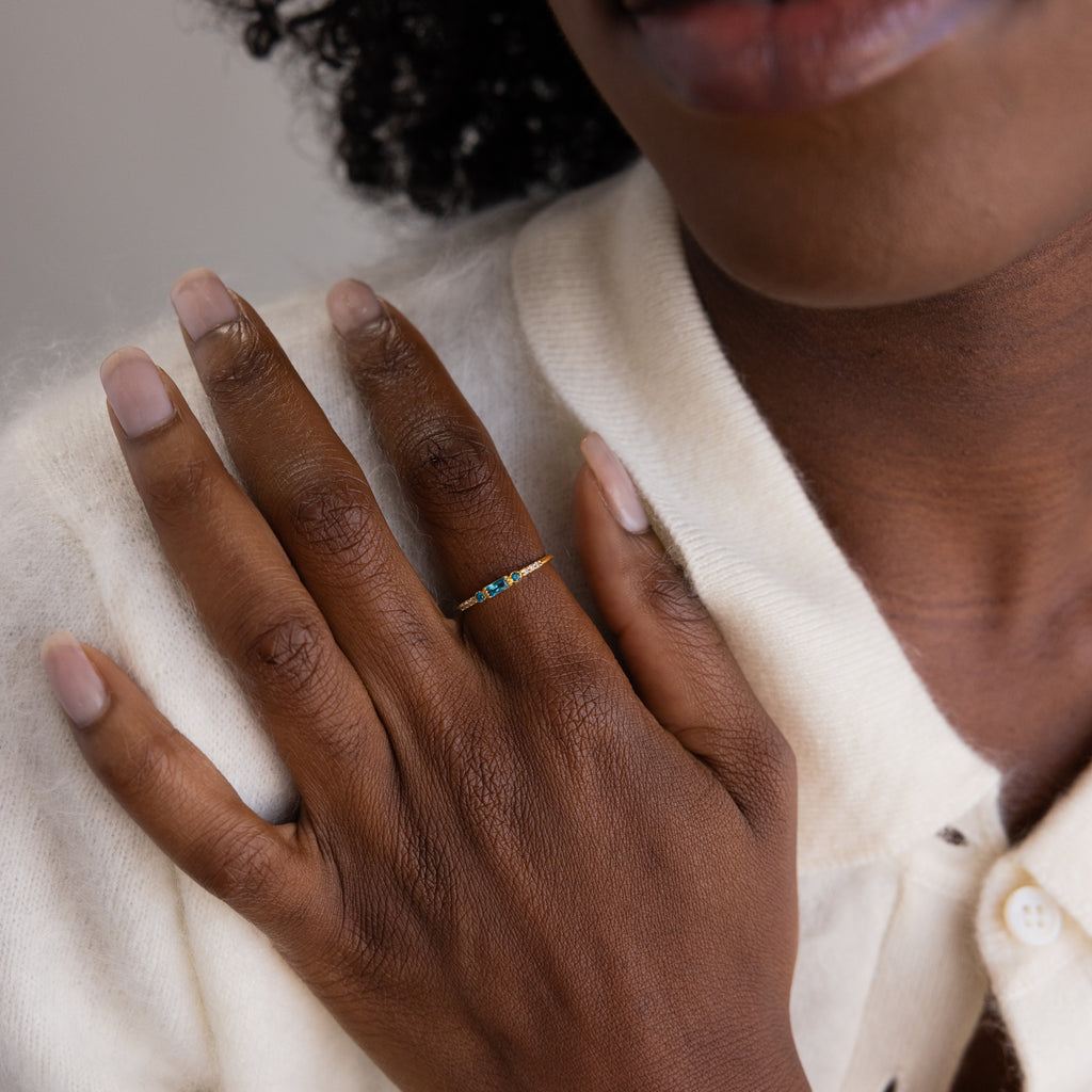A hand placed over the chest, wearing the dainty birthstone ring with a striking blue baguette-cut stone set in a thin gold band.