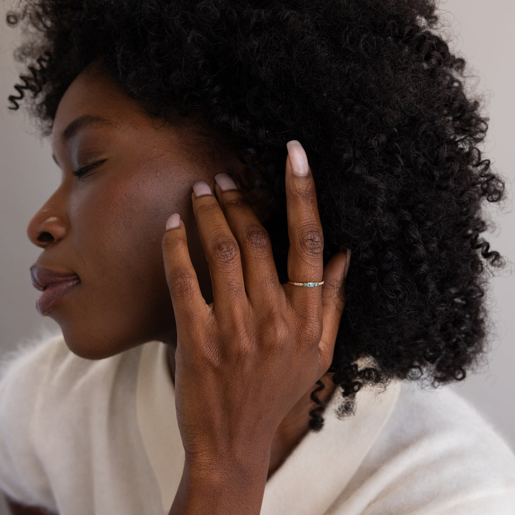 A serene side profile of a woman touching her hair, displaying the dainty gold ring with a brilliant blue gemstone on her ring finger.