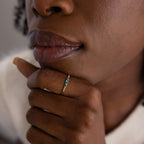 Zoomed-in view of a woman's hand resting near her lips, highlighting a dainty gold birthstone ring with a vivid blue baguette-cut gemstone.