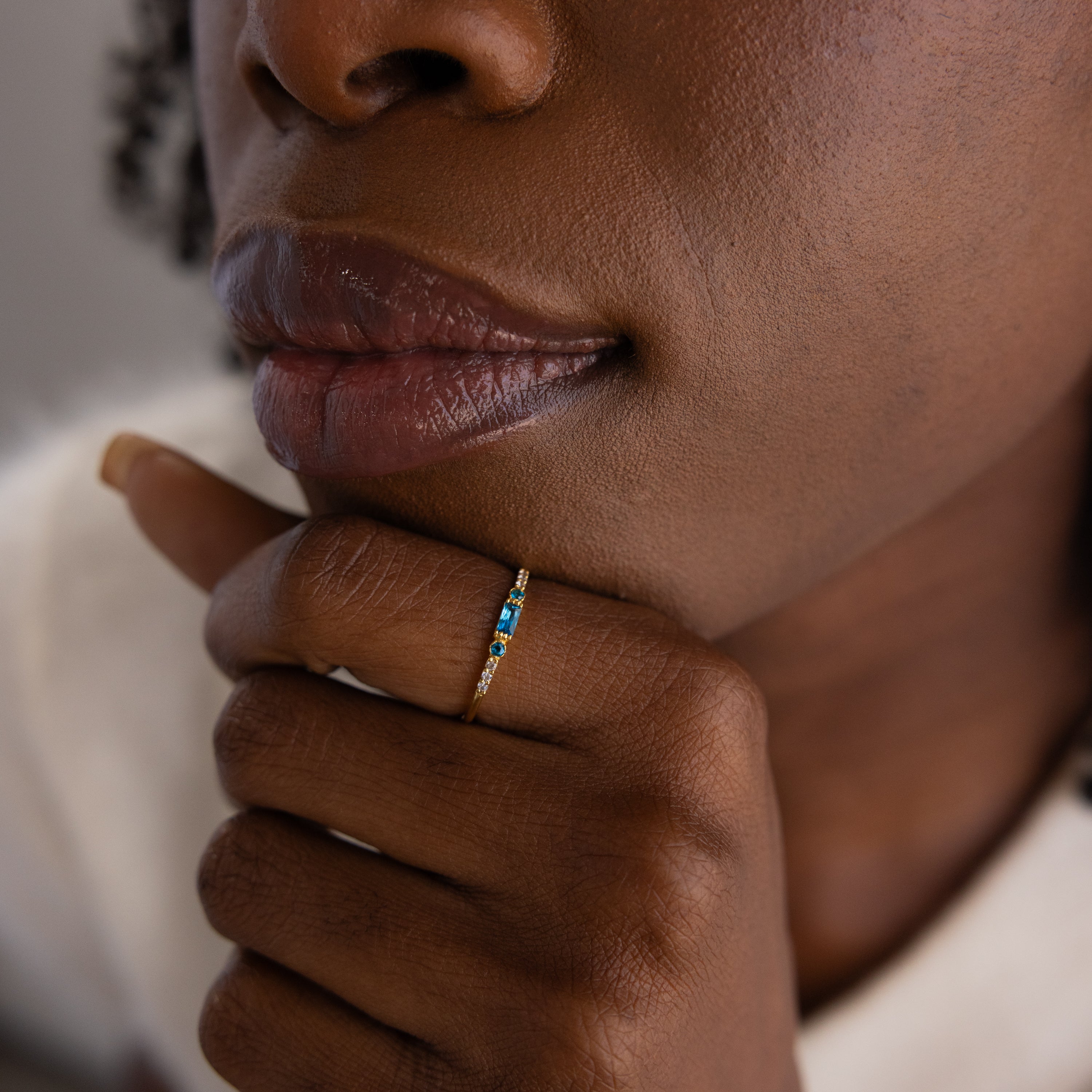 Zoomed-in view of a woman's hand resting near her lips, highlighting a dainty gold birthstone ring with a vivid blue baguette-cut gemstone.