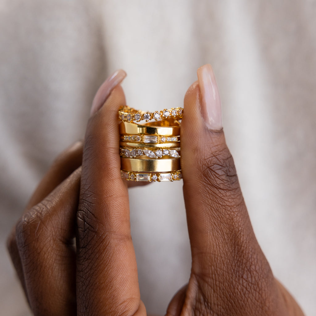 Close-up of hands holding a stack of gold rings, featuring a mix of sparkly designs, including a dainty birthstone ring with red and white stones.