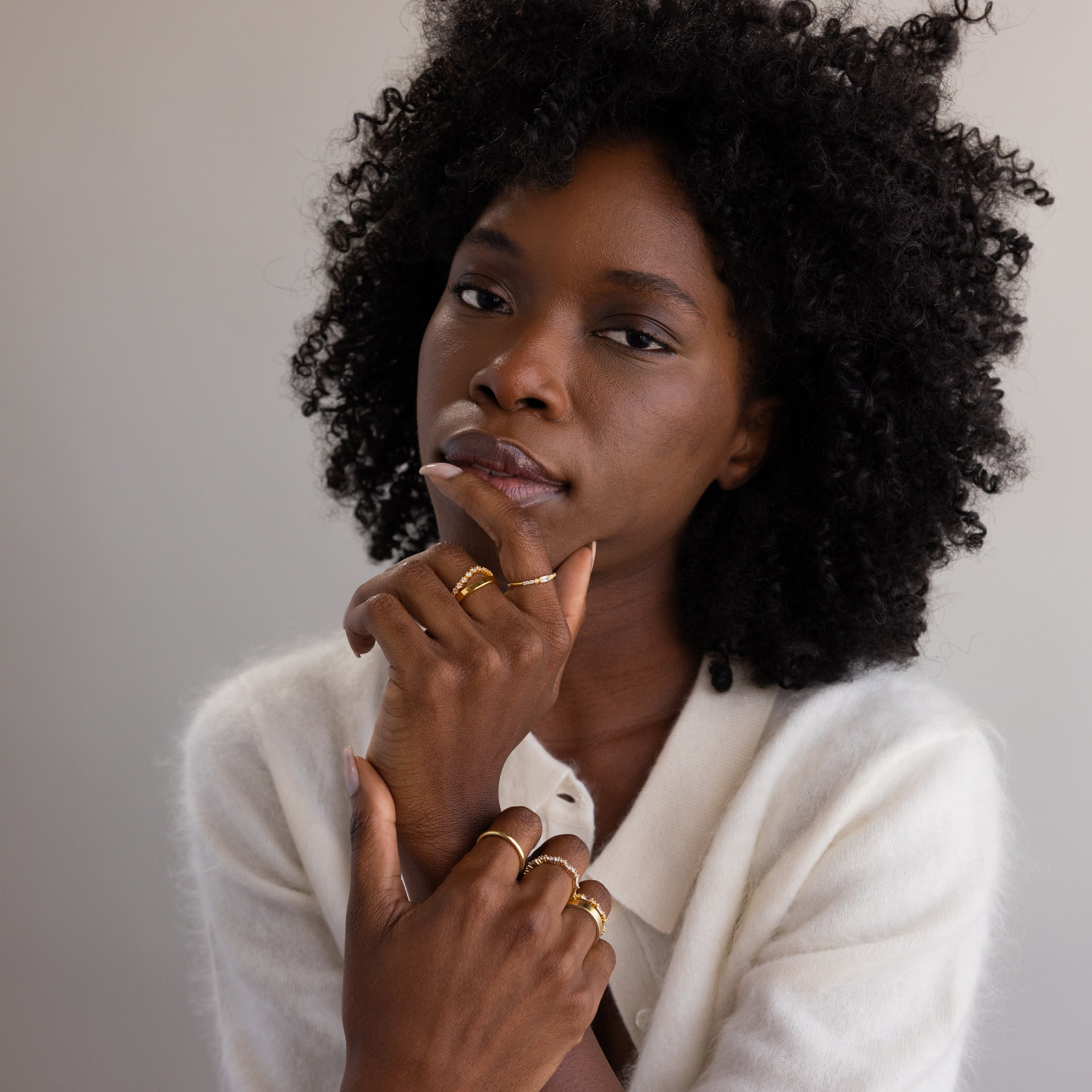 A poised woman rests her chin on her hand, showcasing the dainty gold birthstone ring among several other stacked gold bands, all worn on her fingers with a soft ivory cardigan.