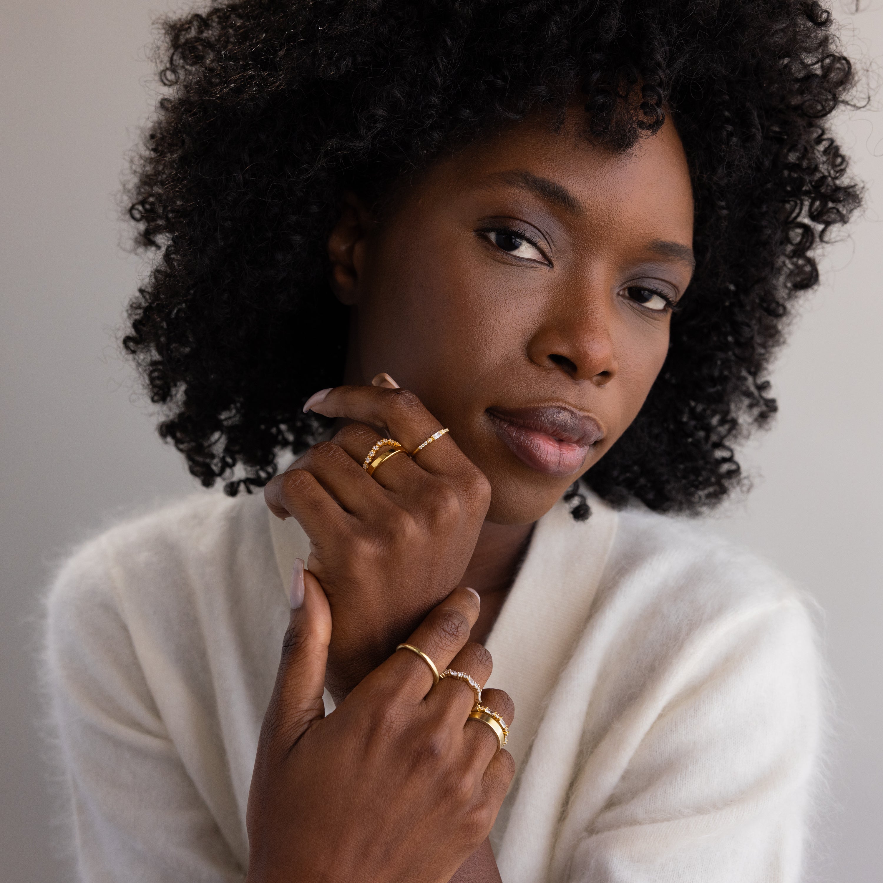 A woman with natural curly hair wears multiple gold rings on her fingers, including the dainty birthstone ring with a red gemstone, styled with a cozy ivory sweater.