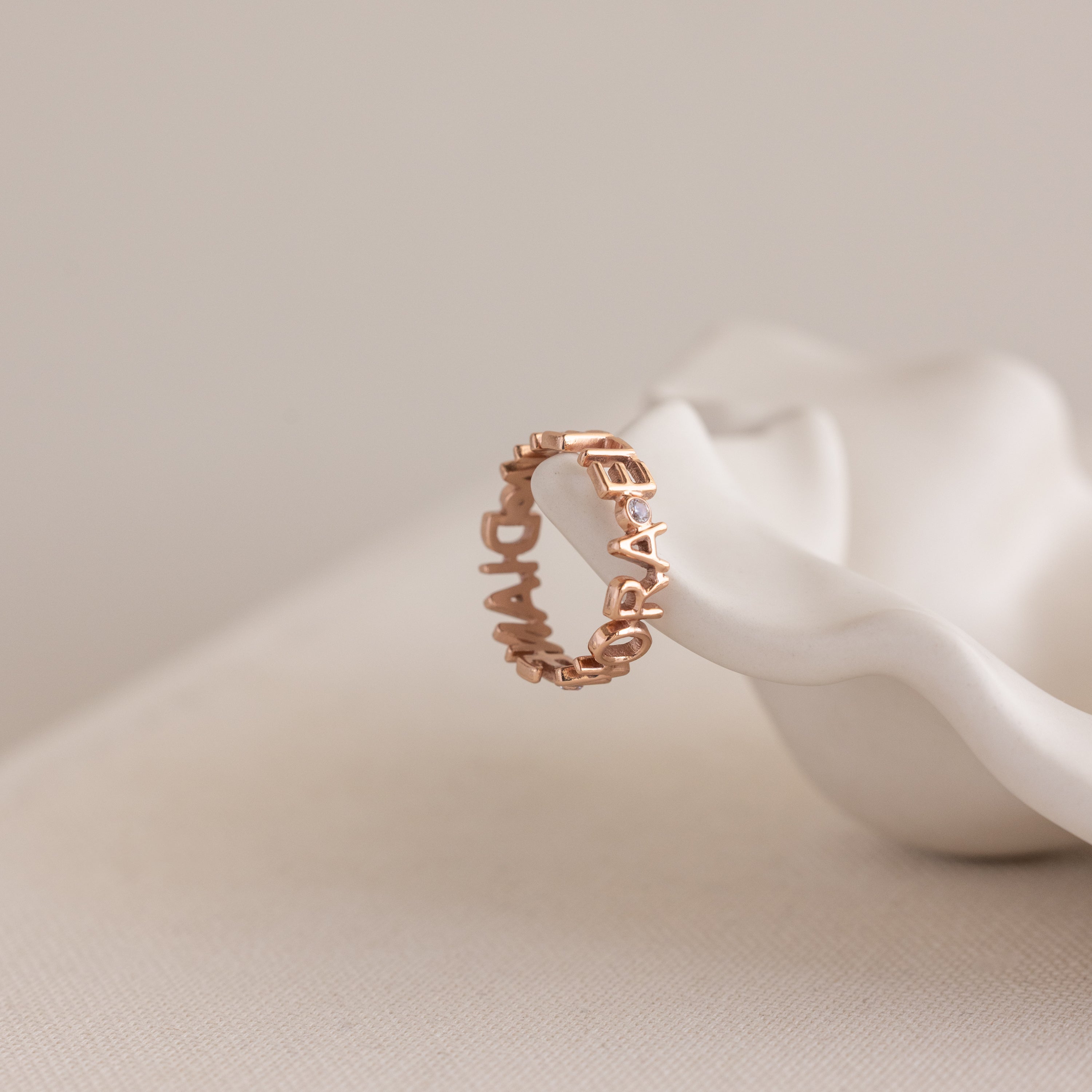 The Multiple Name Eternity Ring in Rose Gold features personalized raised letters and sparkling diamond accents, elegantly displayed on a white sculpted ceramic dish.