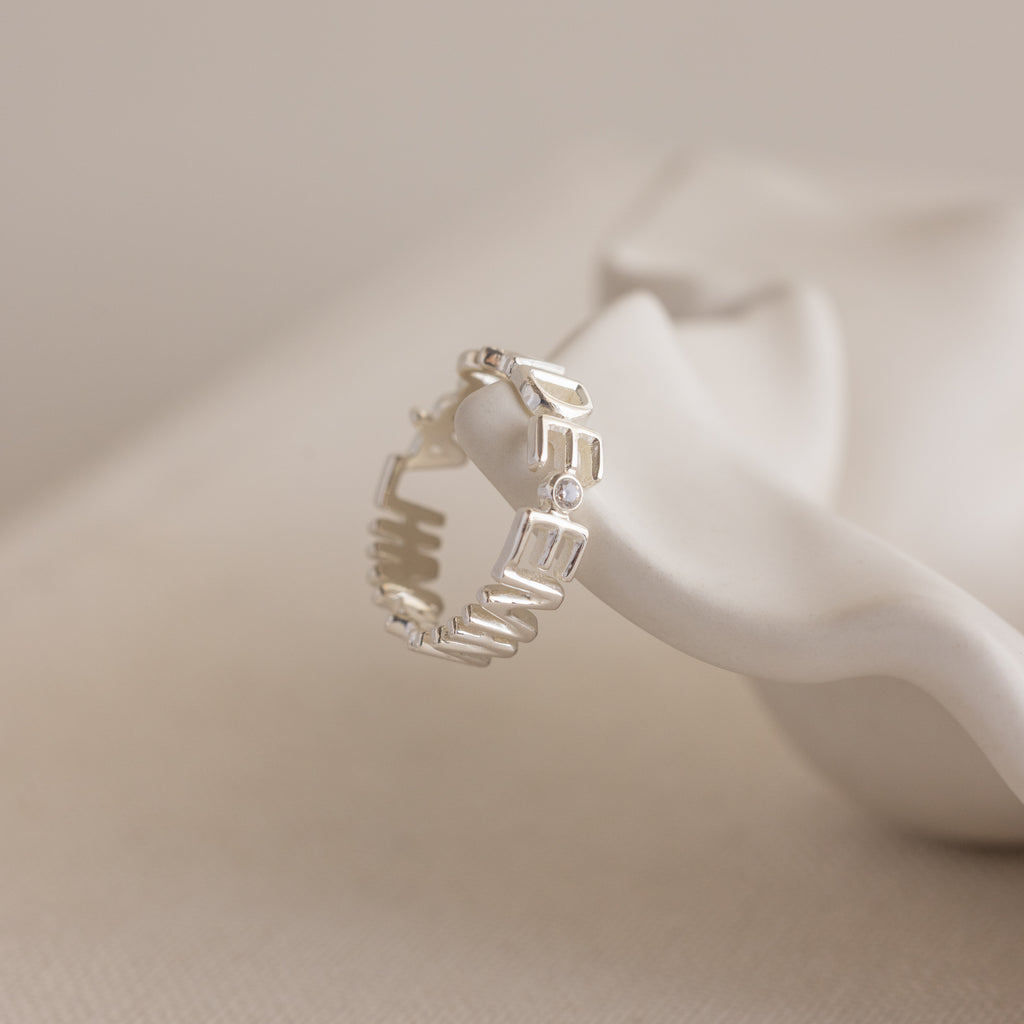 A Multiple Name Eternity Ring in sterling silver, displayed on a white sculptural surface, personalized with the word PEACE.